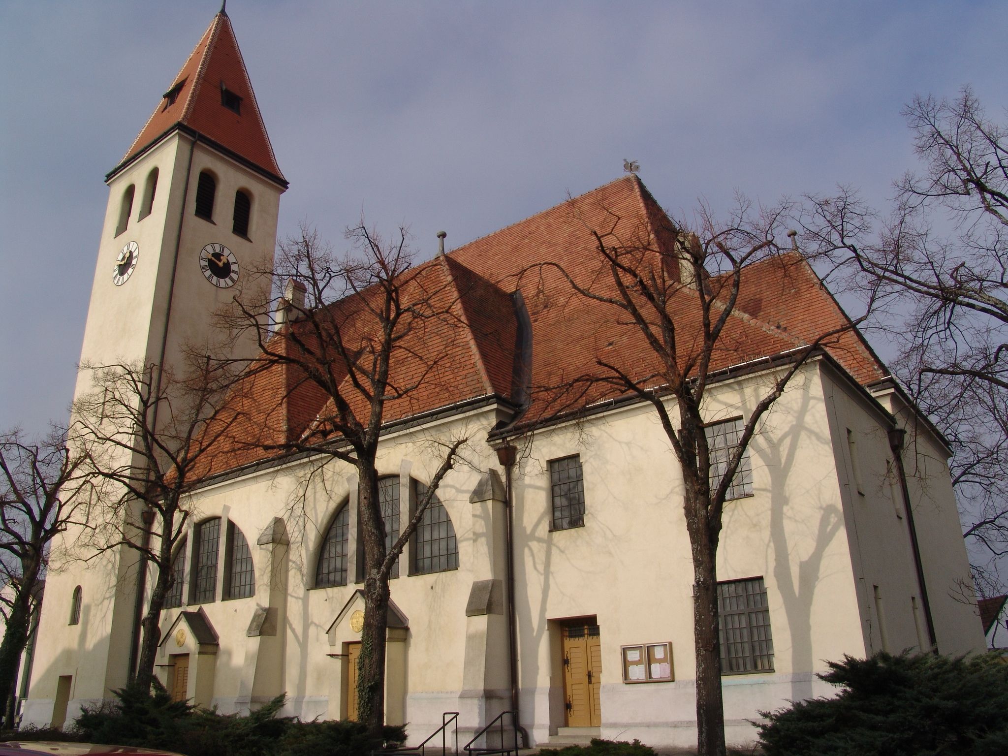 Kostel s věží a červenou taškovou střechou, obklopený holými stromy.