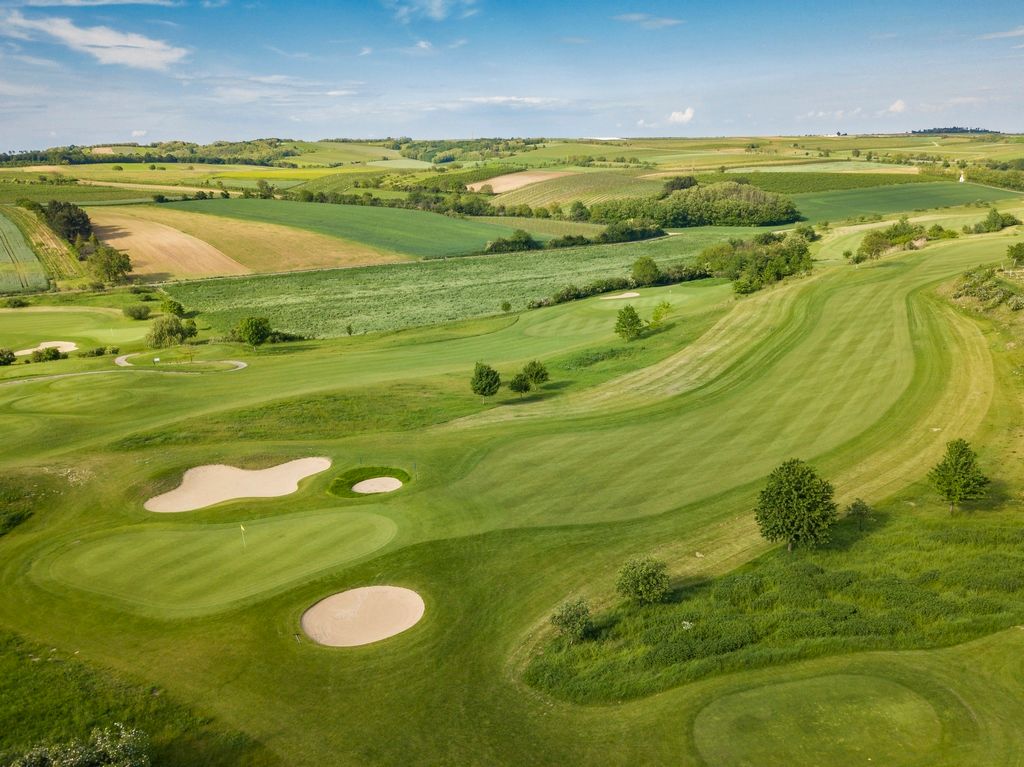 Letecký pohled na golfové hřiště se zelenými fairwayemi a písečnými bunkry, obklopené poli a kopci.
