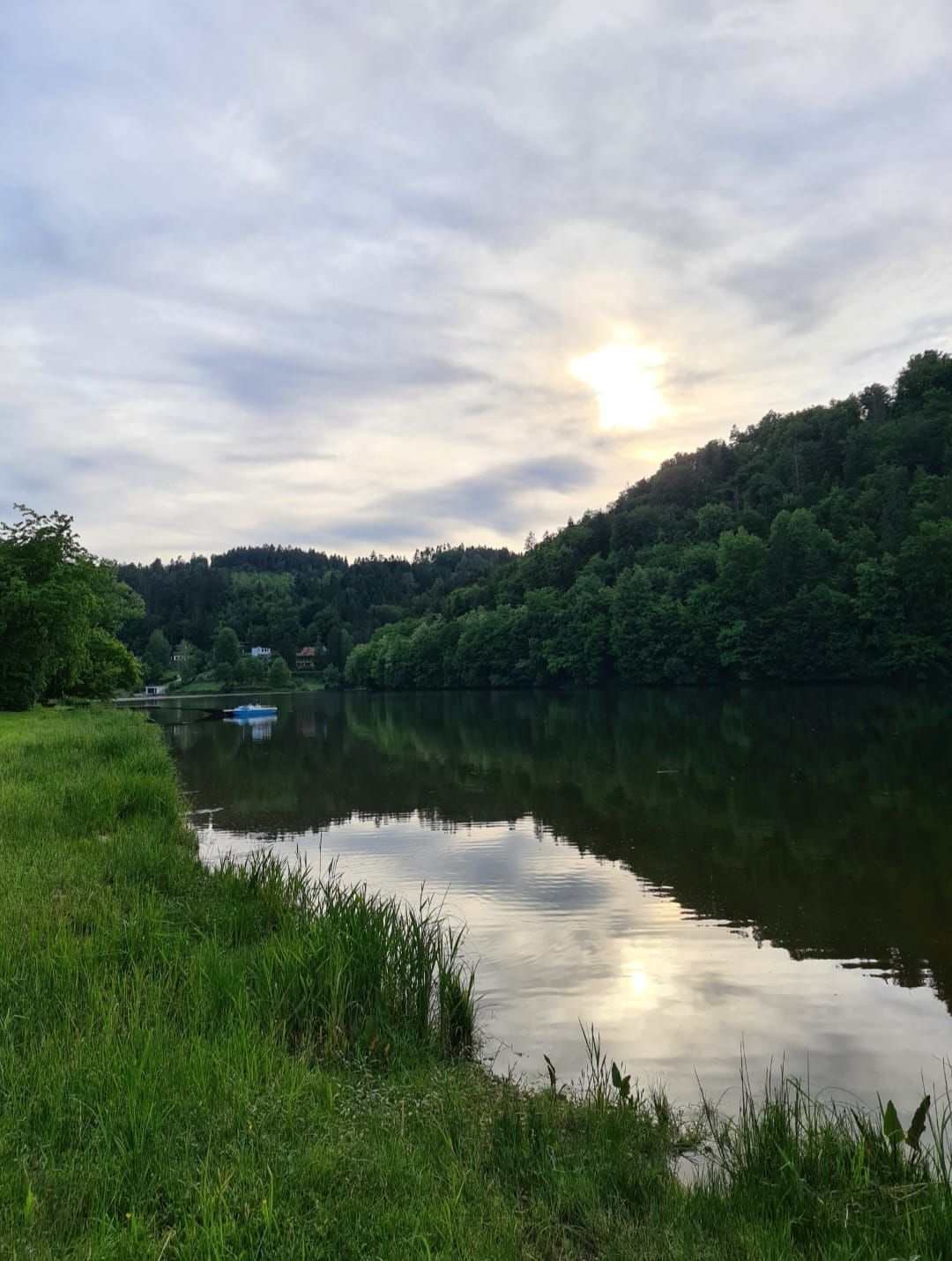 Klidné jezero se zalesněnými kopci v pozadí a mírně zamračeným večerním sluncem.