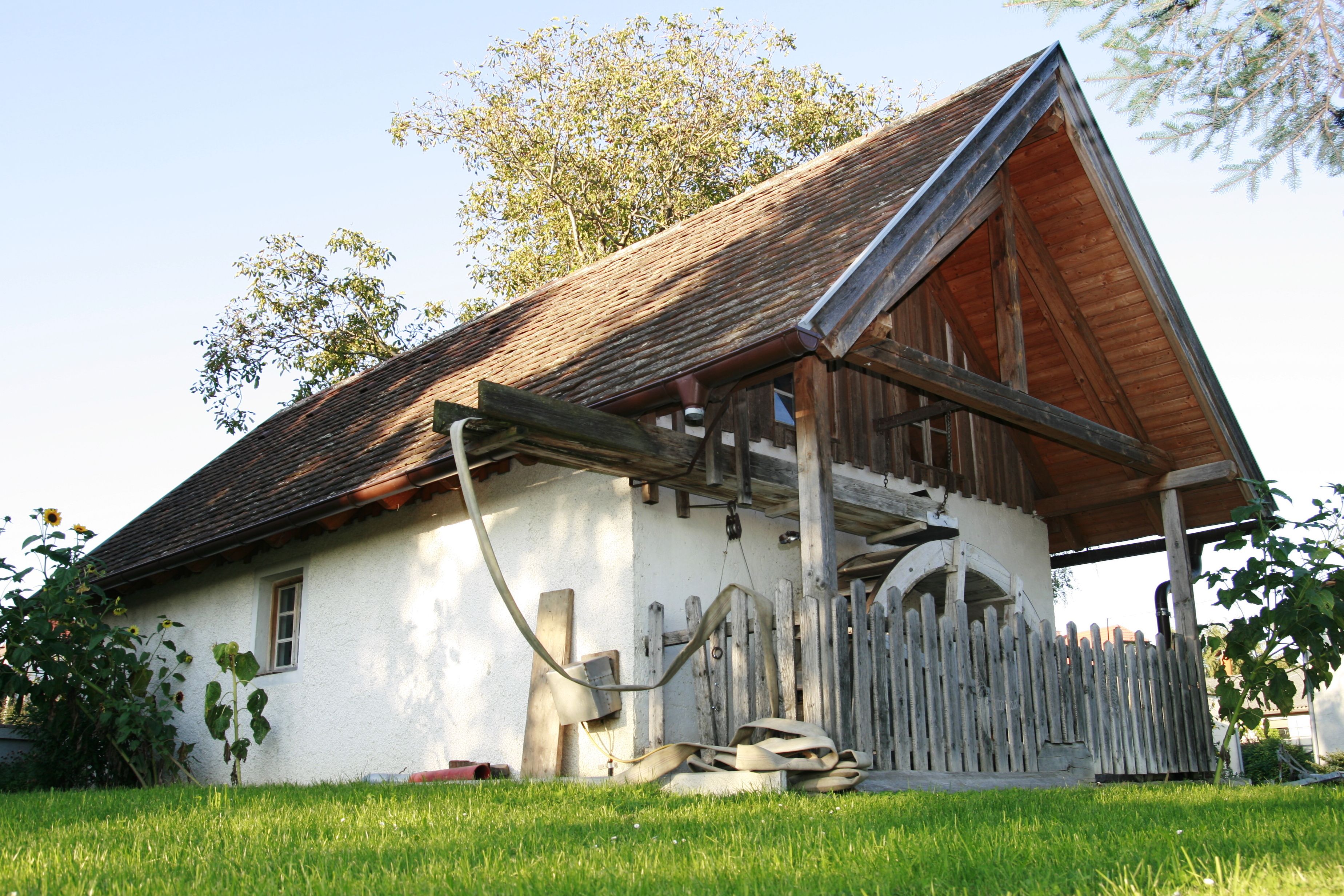Tradiční stavení s dřevěnou střechou a plotem, obklopené zelenou loukou a stromy.