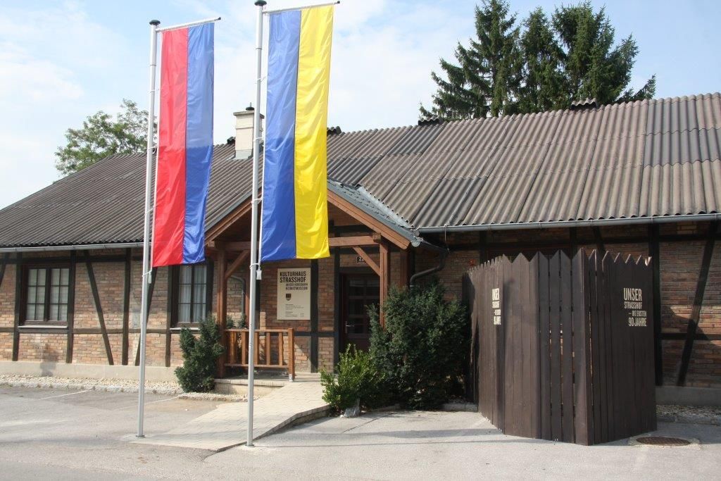 Vchod do muzea místní historie Strasshof se dvěma stožáry a dřevěným plotem.