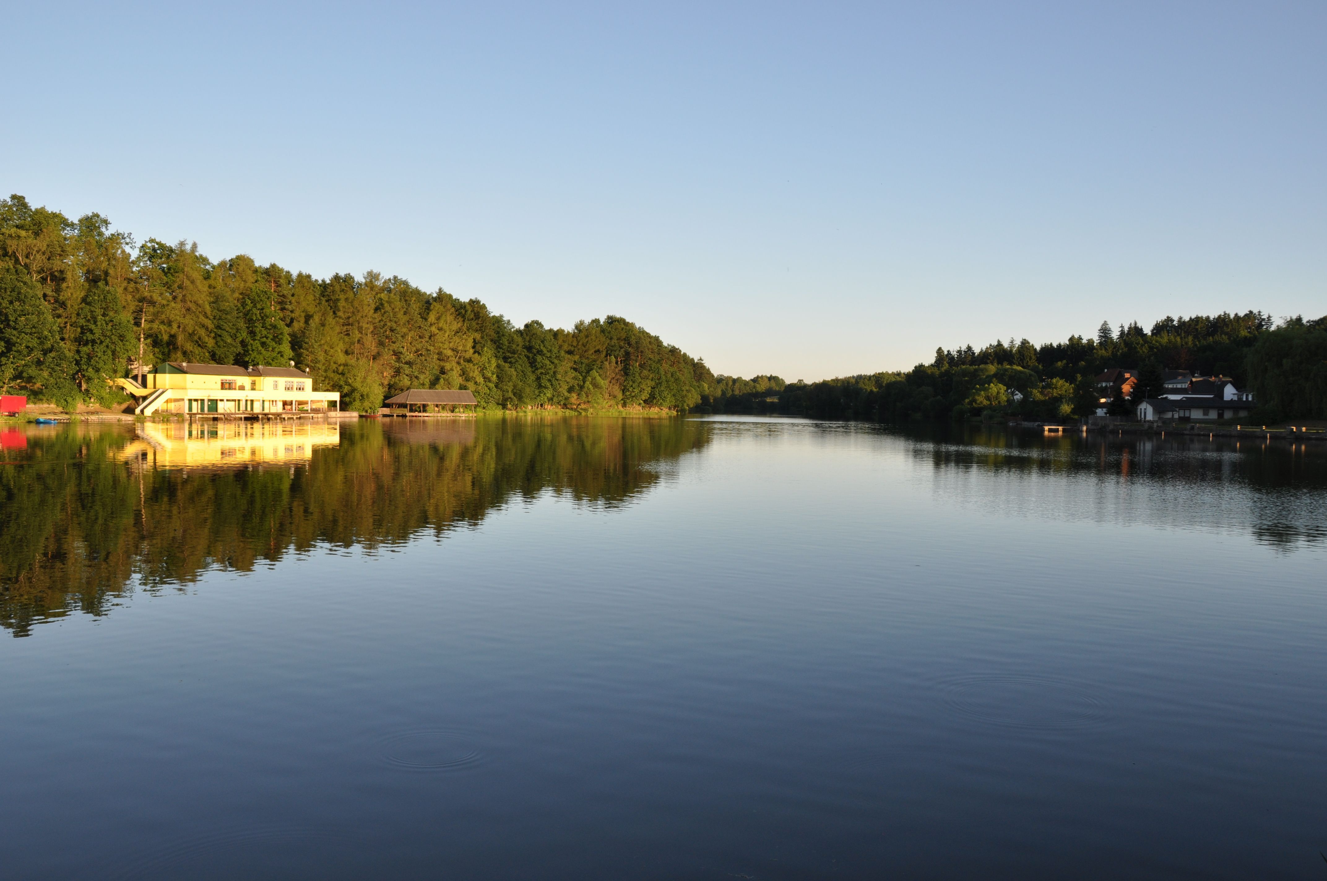 Klidné jezero s budovou na břehu, obklopené stromy, při západu slunce.