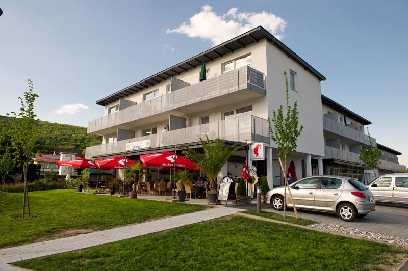 Pohled zvenčí na moderní budovu s kavárnou a červenými slunečníky.