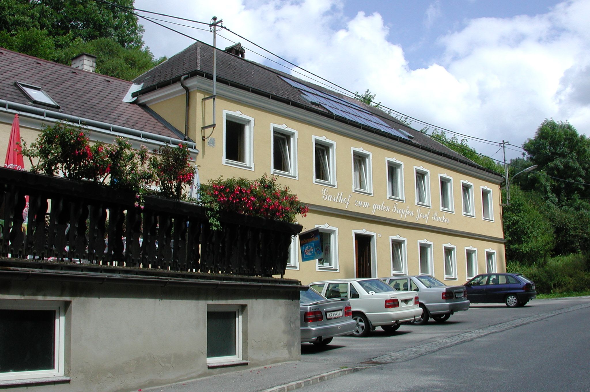 Žlutá budova s nápisem "Gasthof zum guten Tropfen Josef Rucker", před ní zaparkovaná auta, v pozadí modrá obloha.