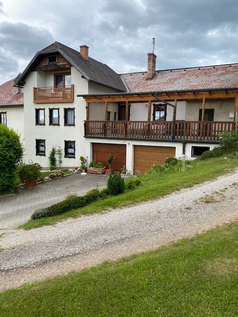 Dvoupodlažní dům s balkonem a garáží, obklopený zelenou krajinou a zamračenou oblohou.