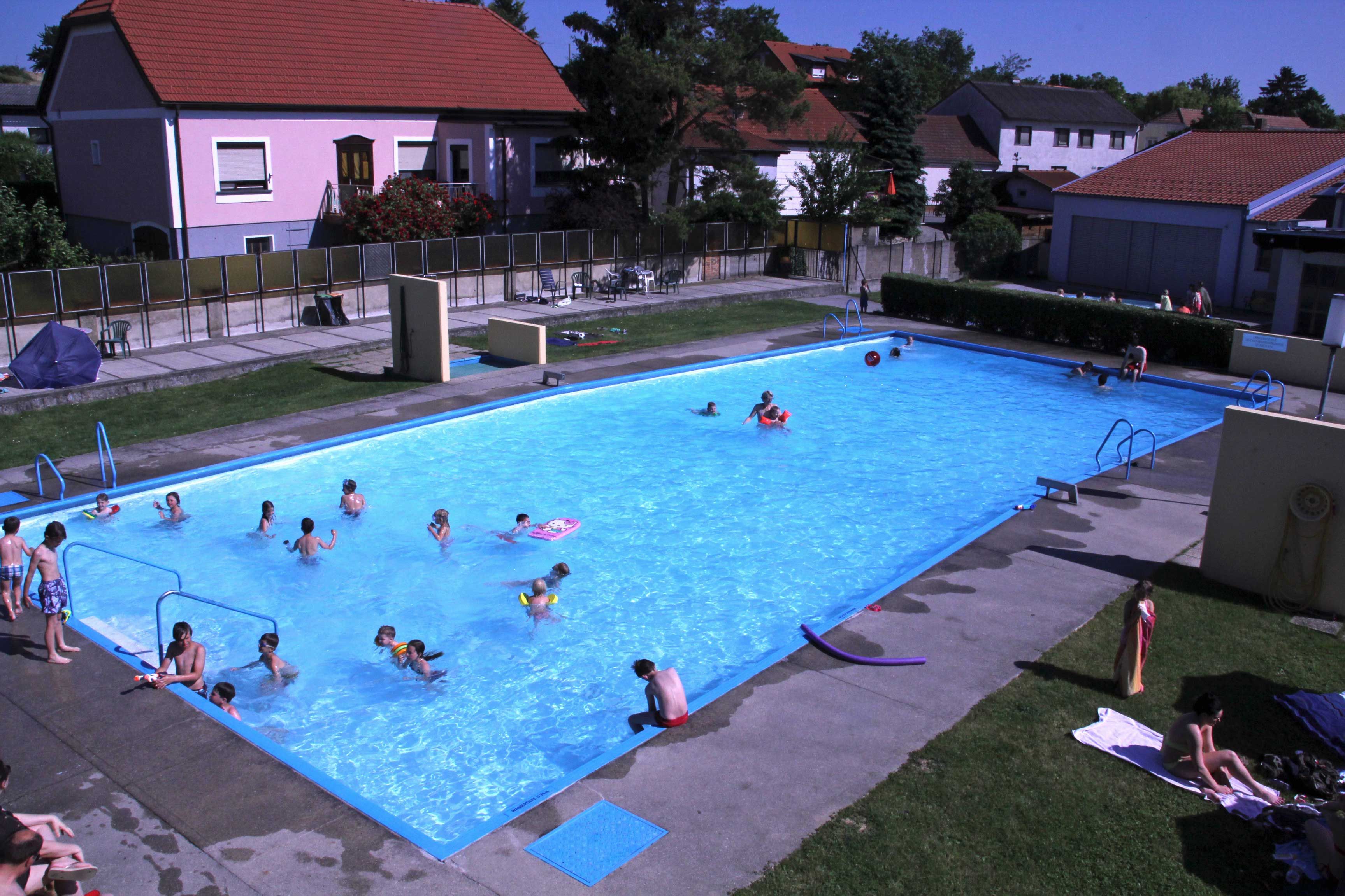 Venkovní bazén s několika lidmi ve vodě a na trávníku na opalování.