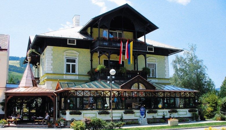 Tradiční budova s prosklenou verandou a rakouskou vlajkou, obklopená rostlinami.