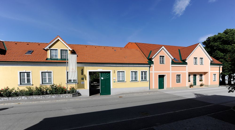 Dlouhý jednopatrový dům se žlutorůžovou fasádou, zelenými okenicemi a červenou taškovou střechou pod modrou oblohou.