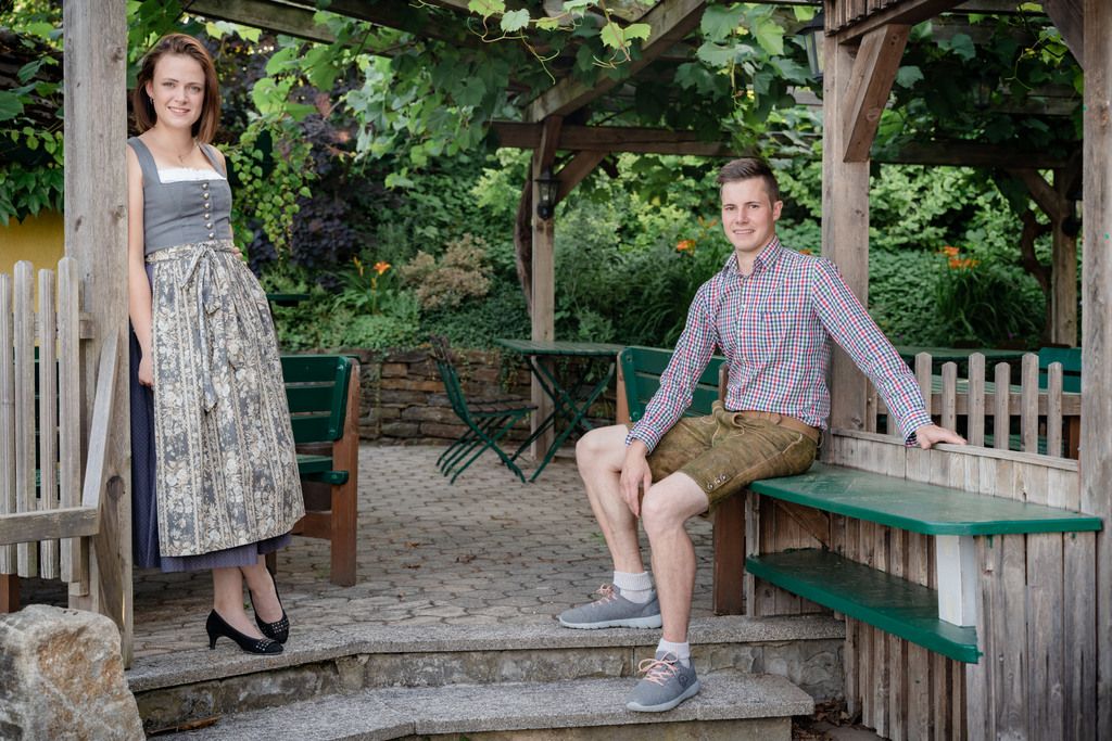 Muž v lederhosen a žena v dirndlu pózují na zahradě s dřevěnými konstrukcemi.