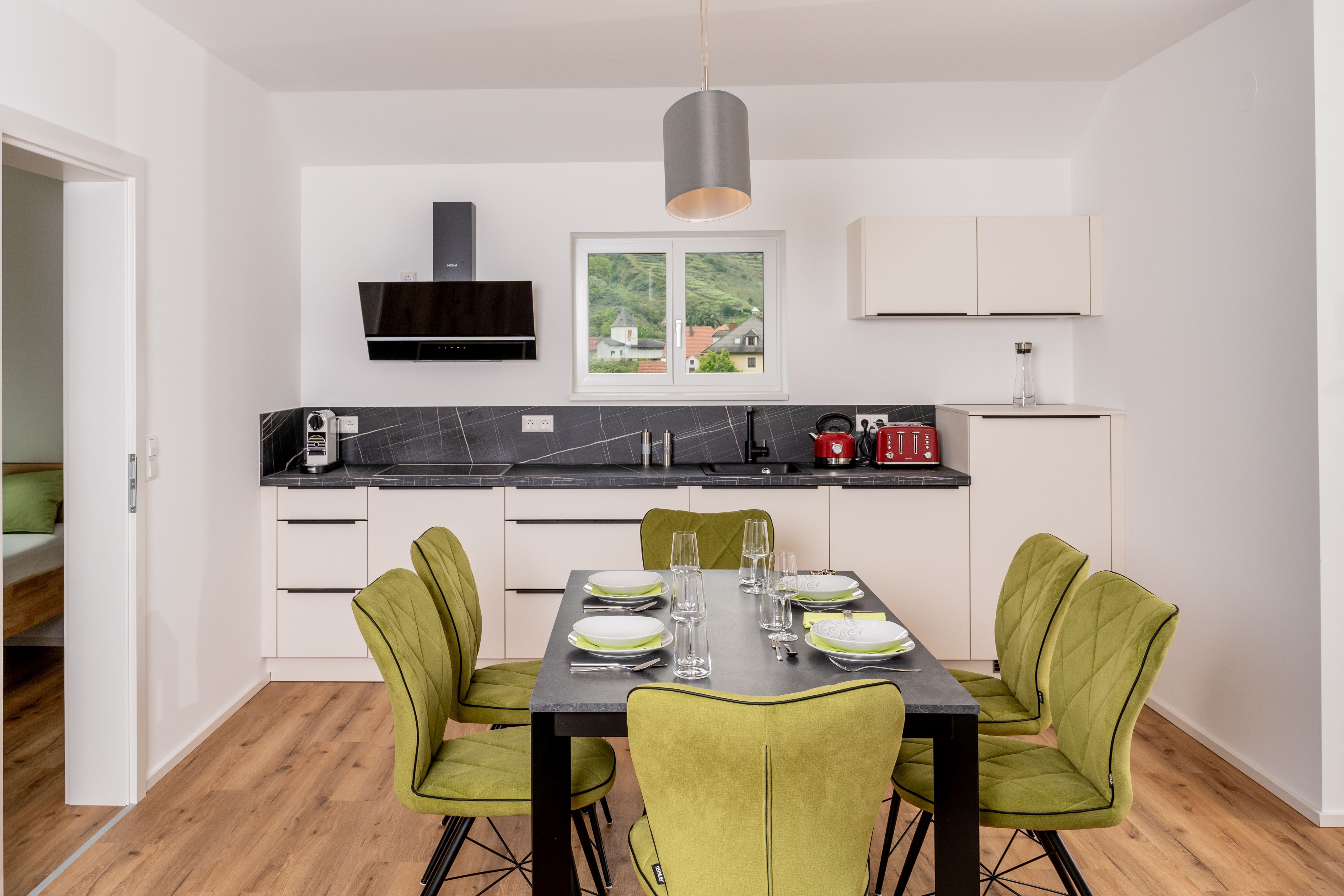 Moderní kuchyně s jídelním stolem a zelenými židlemi, okno s výhledem na kopce.