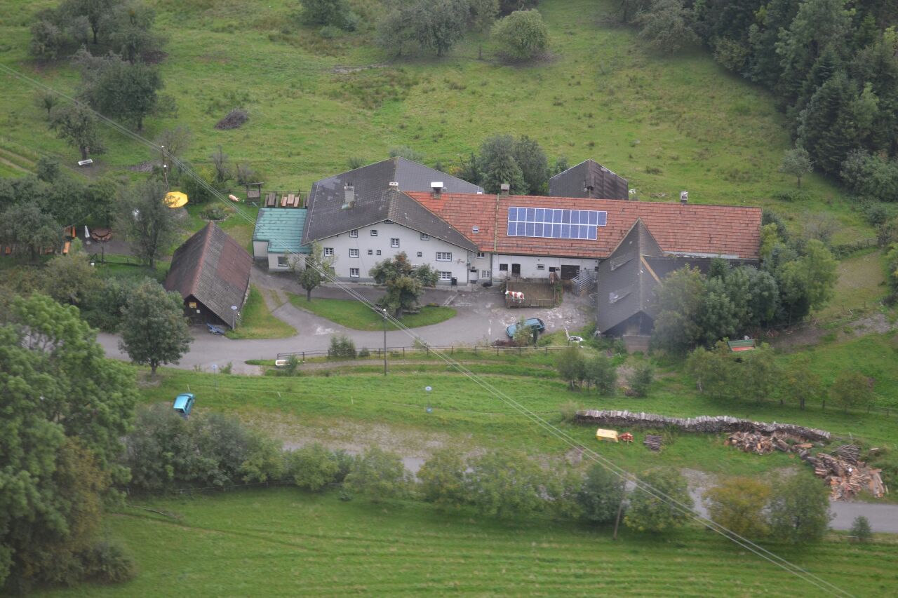 Letecký pohled na farmu se solárními panely na střeše, obklopenou zelenými poli a stromy.