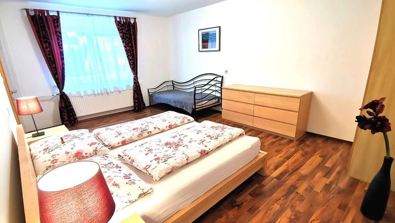 Ložnice s manželskou postelí, jednolůžkem, komodou a květinami ve váze.