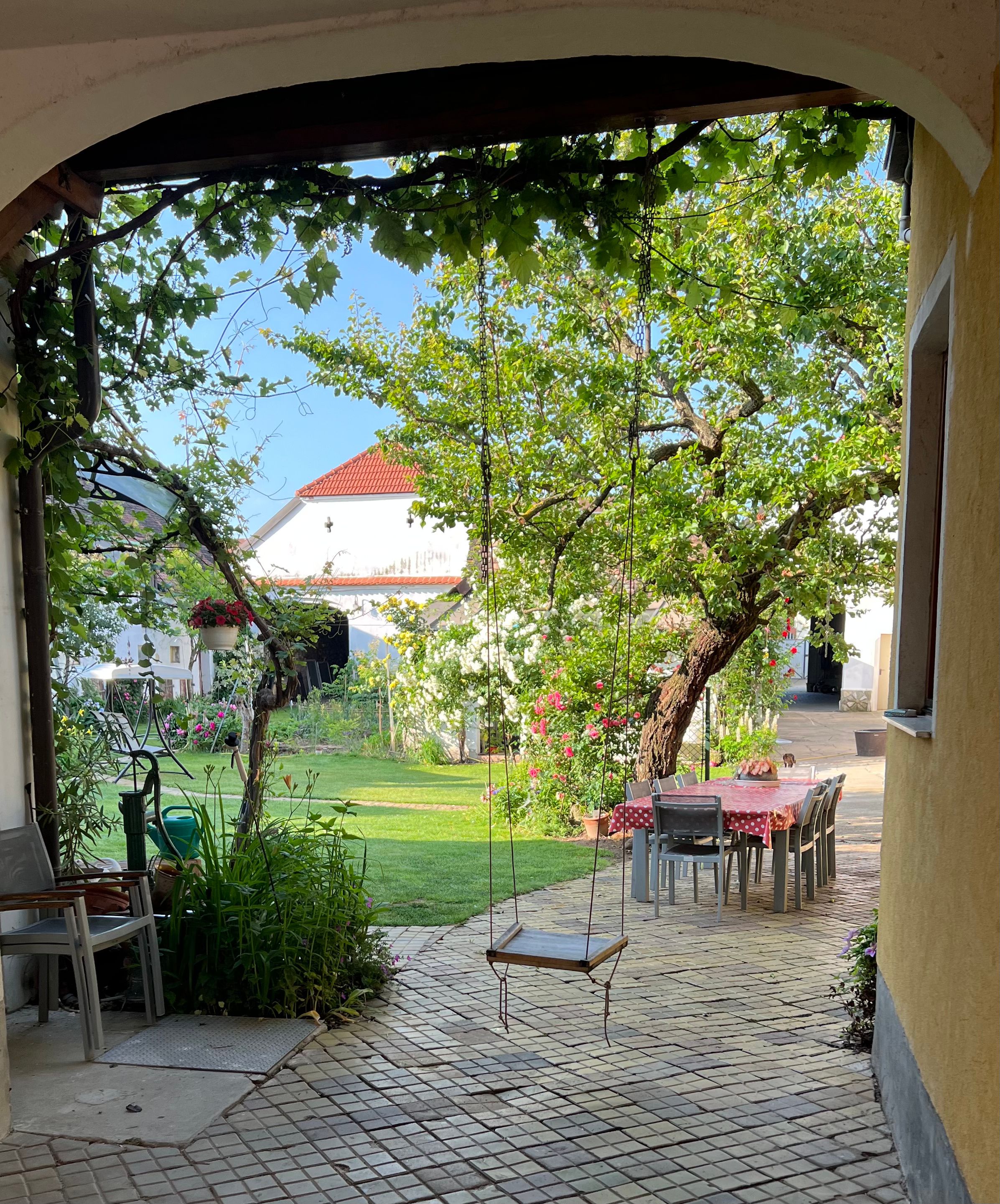 Idylická zahrada s houpačkou, stolem se židlemi a kvetoucími rostlinami.