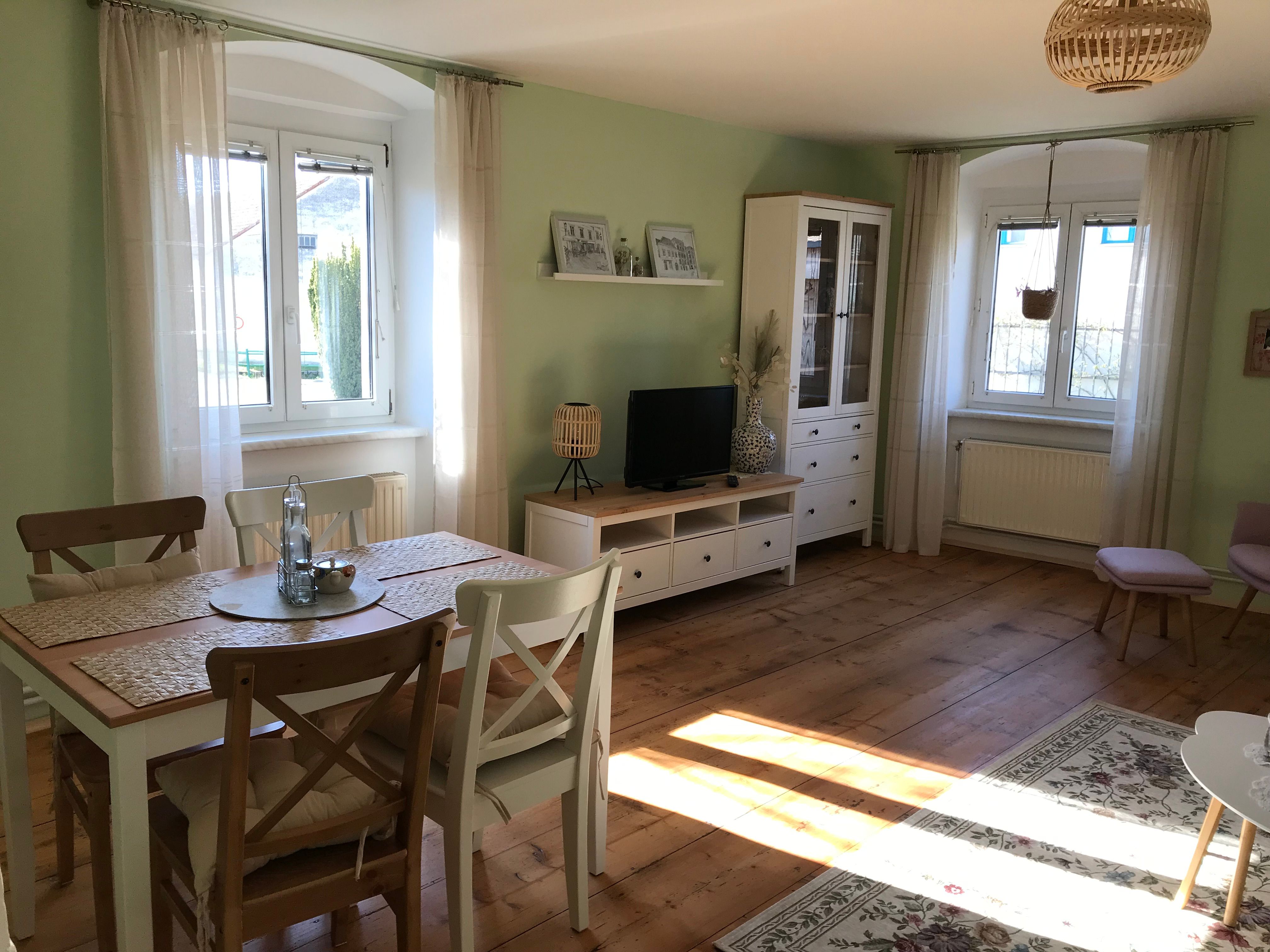 Světlý pokoj s jídelním stolem, židlemi, TV skříňkou a velkými okny.