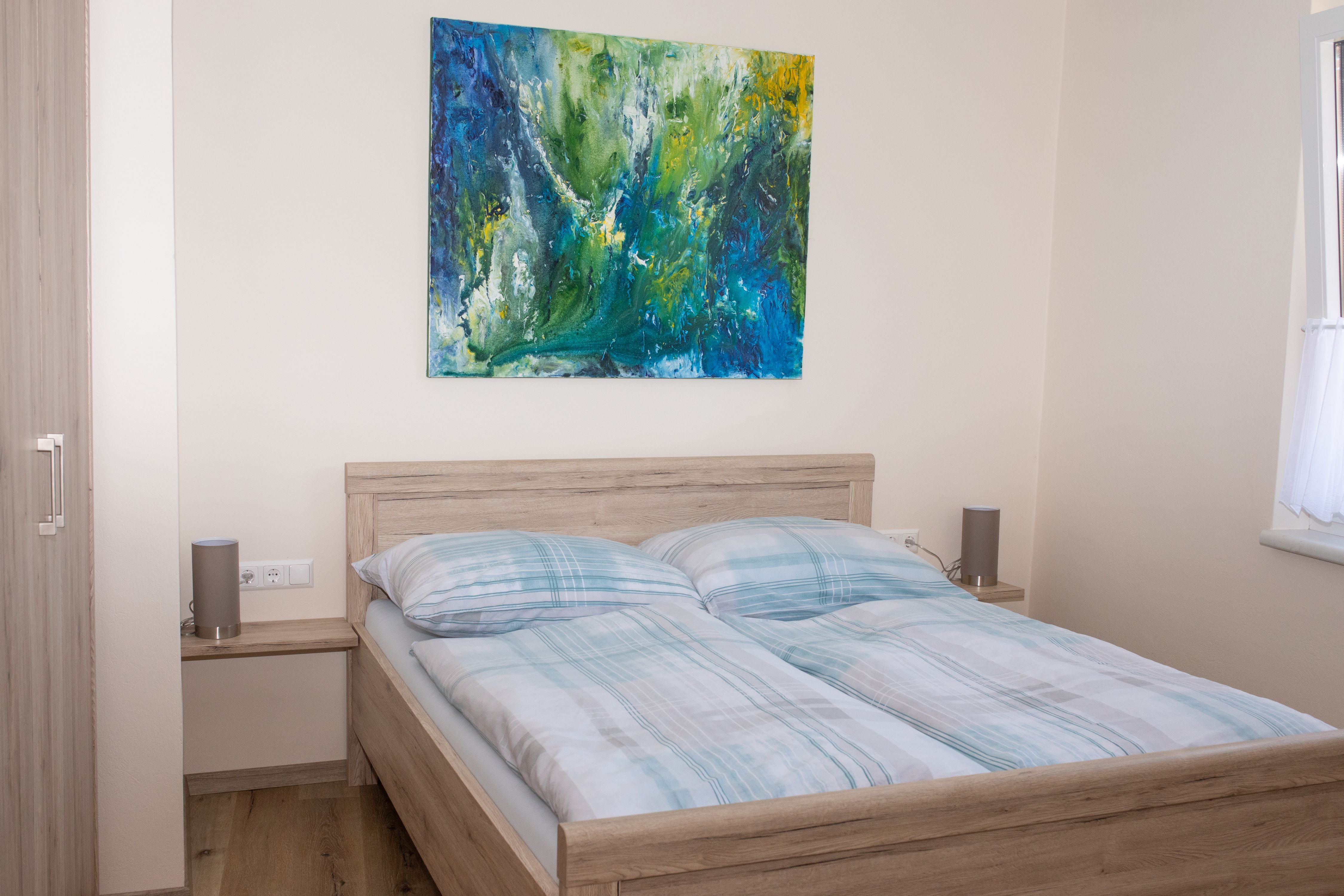Ložnice s manželskou postelí, abstraktní malbou na stěně a oknem se závěsem.