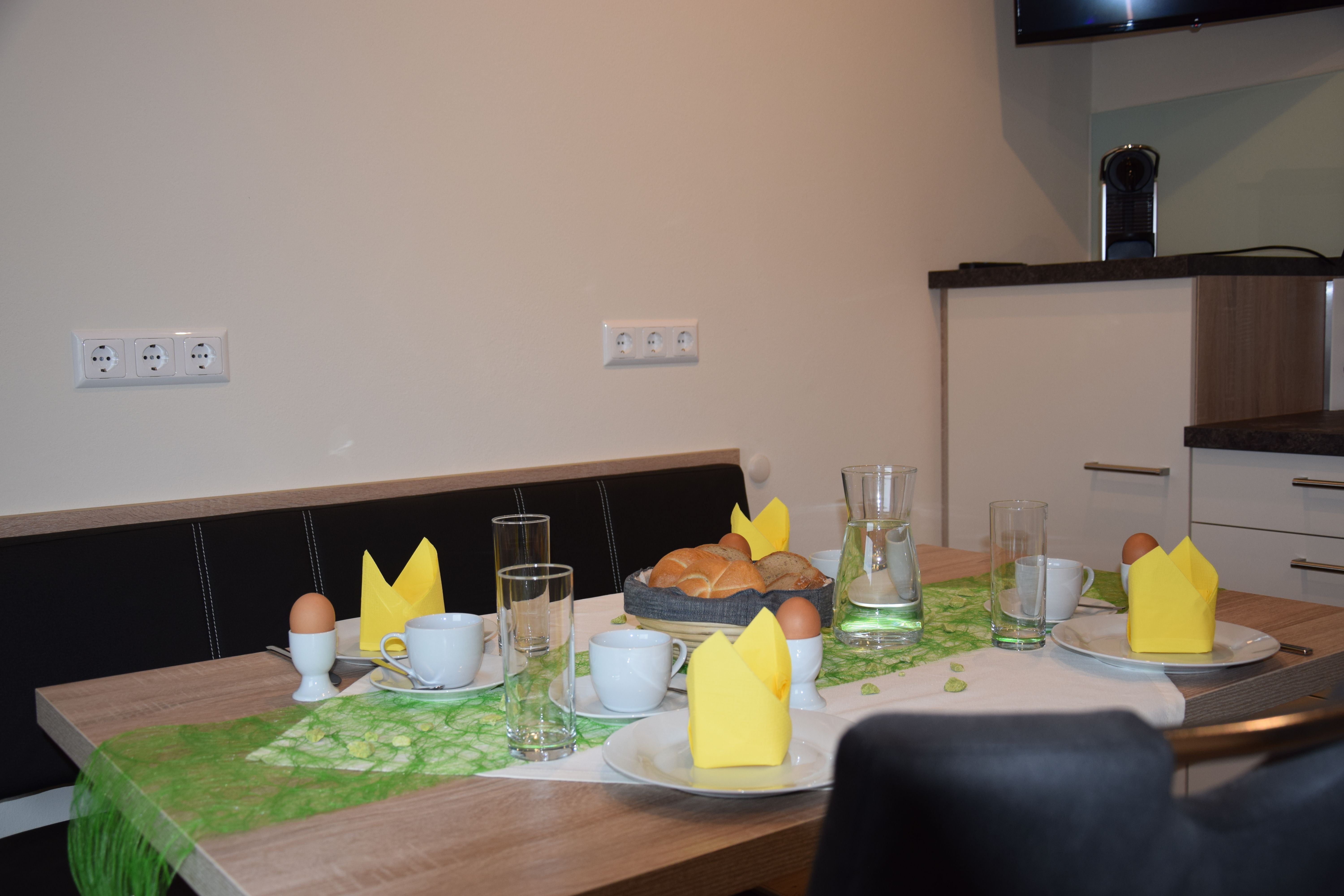 Snídaňový stůl s rohlíky, vejci, šálky a žlutými ubrousky.