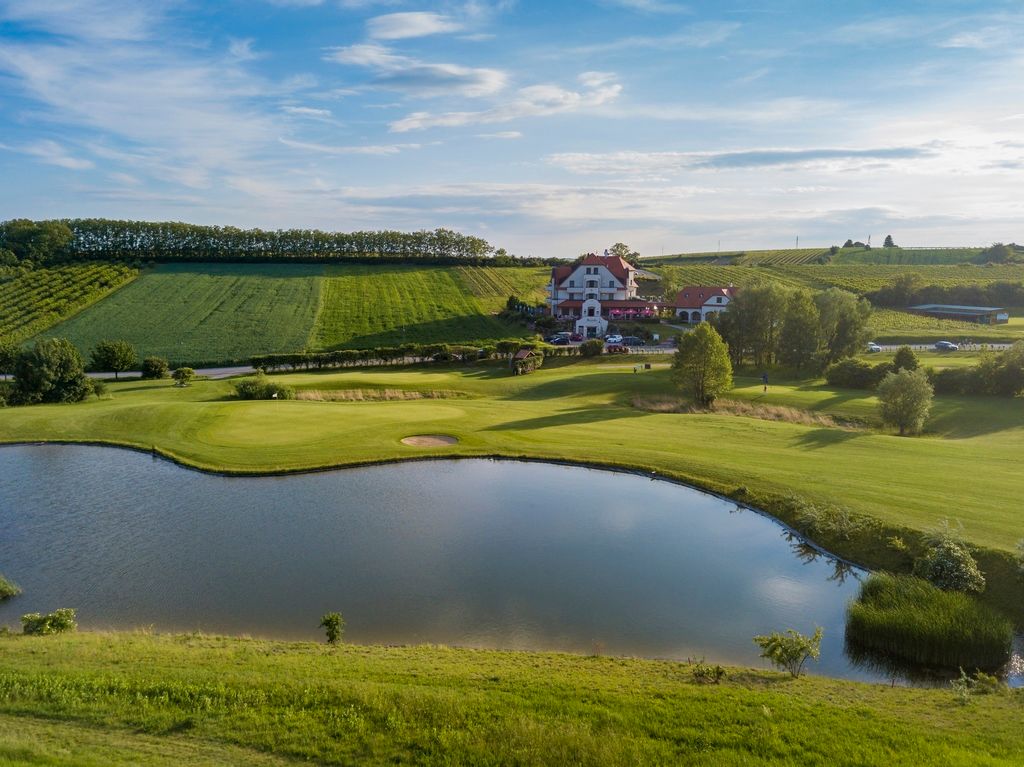 Záběr golfového hřiště s rybníkem a klubovnou v pozadí.