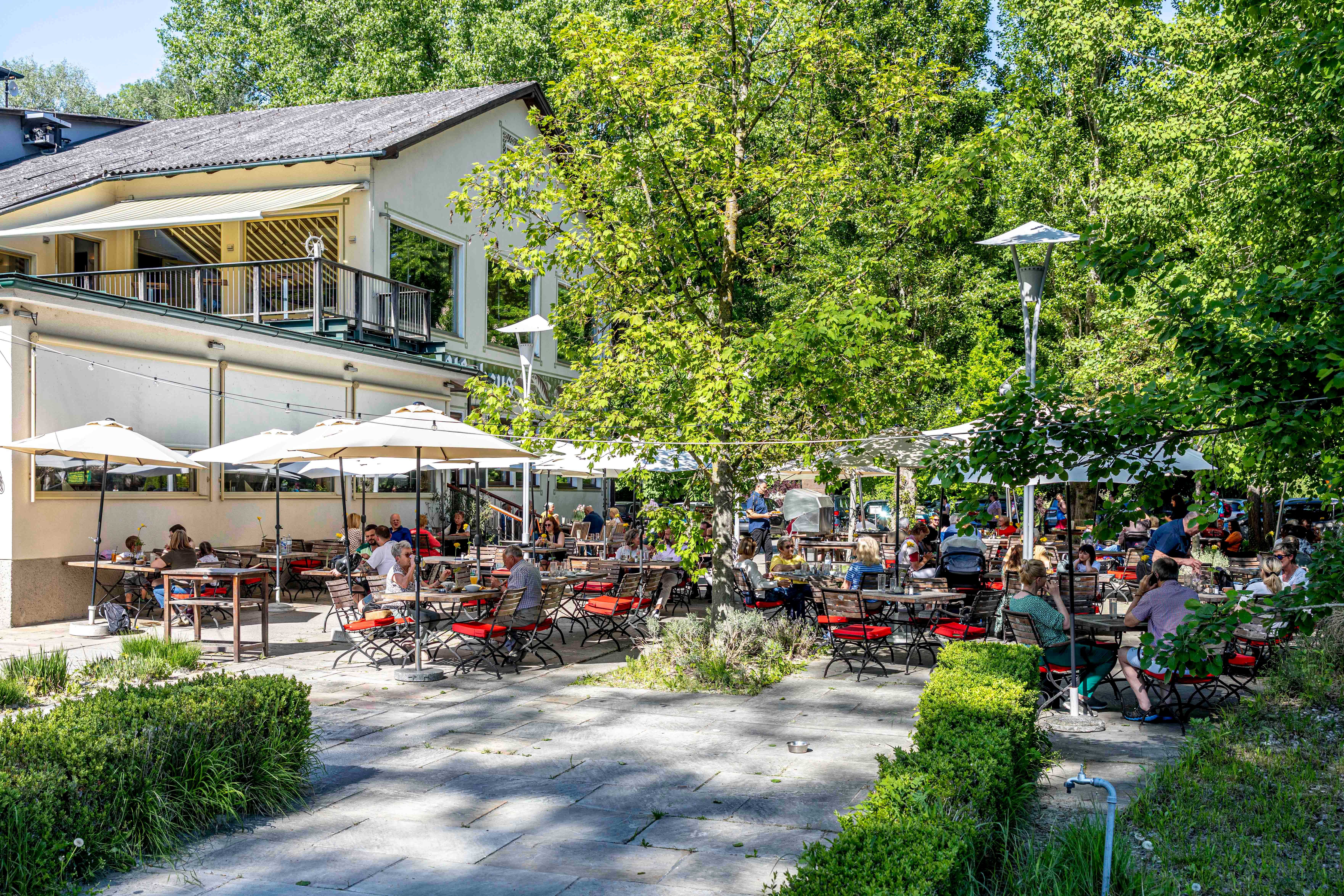 Zahrada pro hosty se spoustou lidí sedících u stolků pod slunečníky, obklopená stromy a budovou v pozadí.