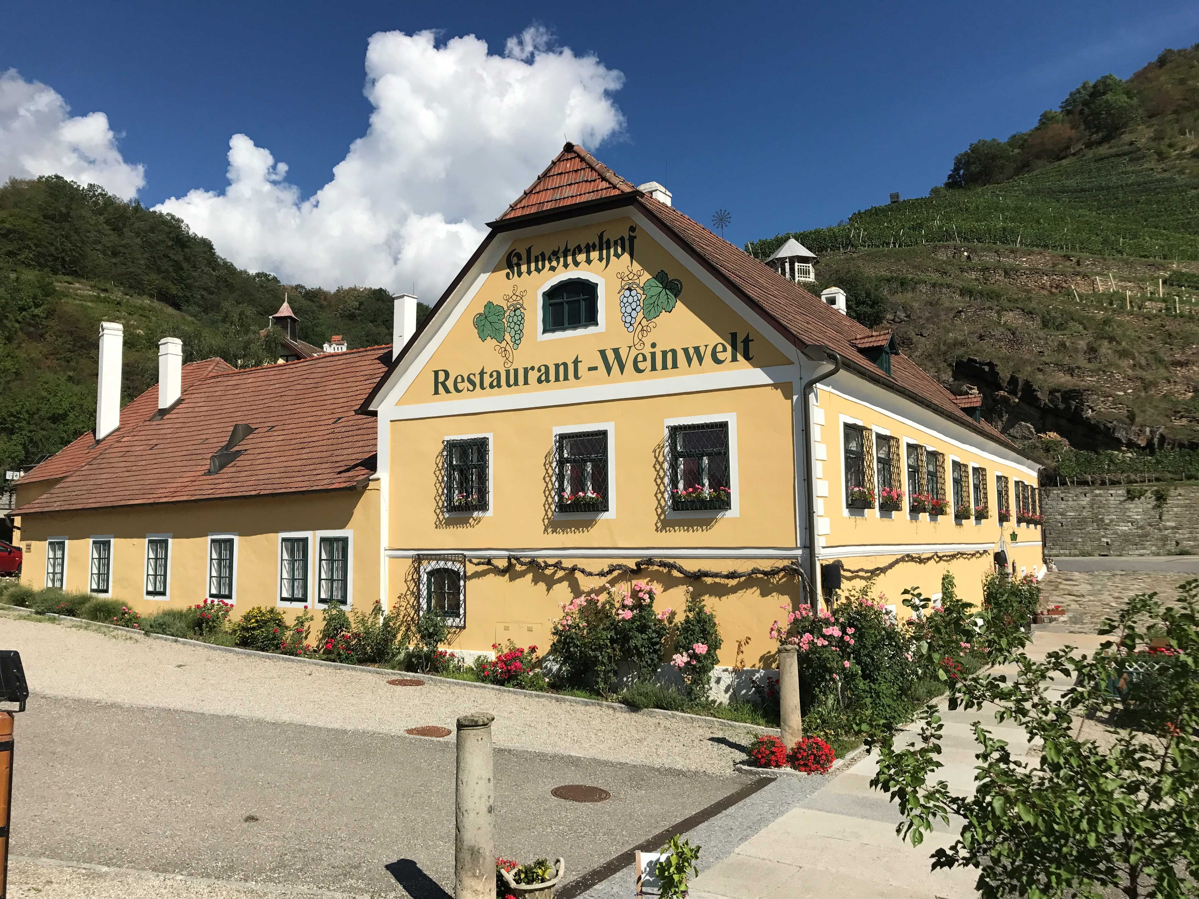 Pohled zvenčí na žlutou budovu s nápisem "Klosterhof Restaurant-Weinwelt", obklopenou květinami a vinicemi.