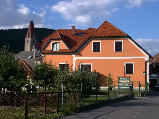Oranžově zbarvený dům s červenými střešními taškami a kostelní věží v pozadí.