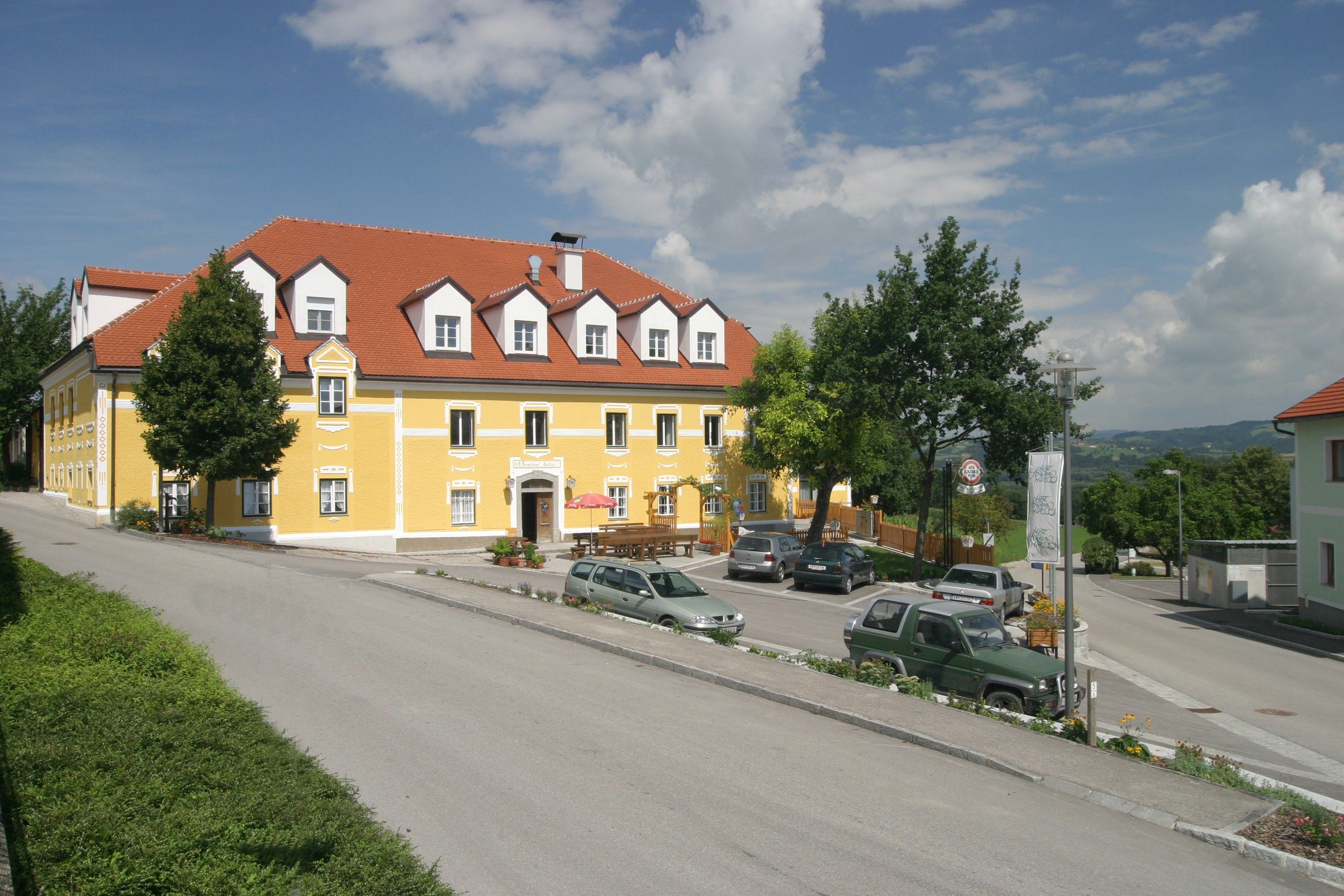 Žlutá budova s červenou střechou, obklopená auty a stromy, pod modrou oblohou.