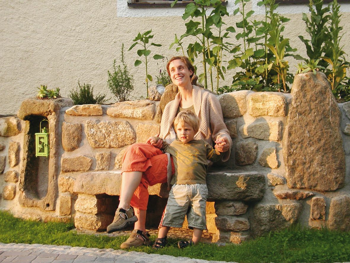 Žena a dítě sedí na kamenné lavičce před skalkou porostlou rostlinami.