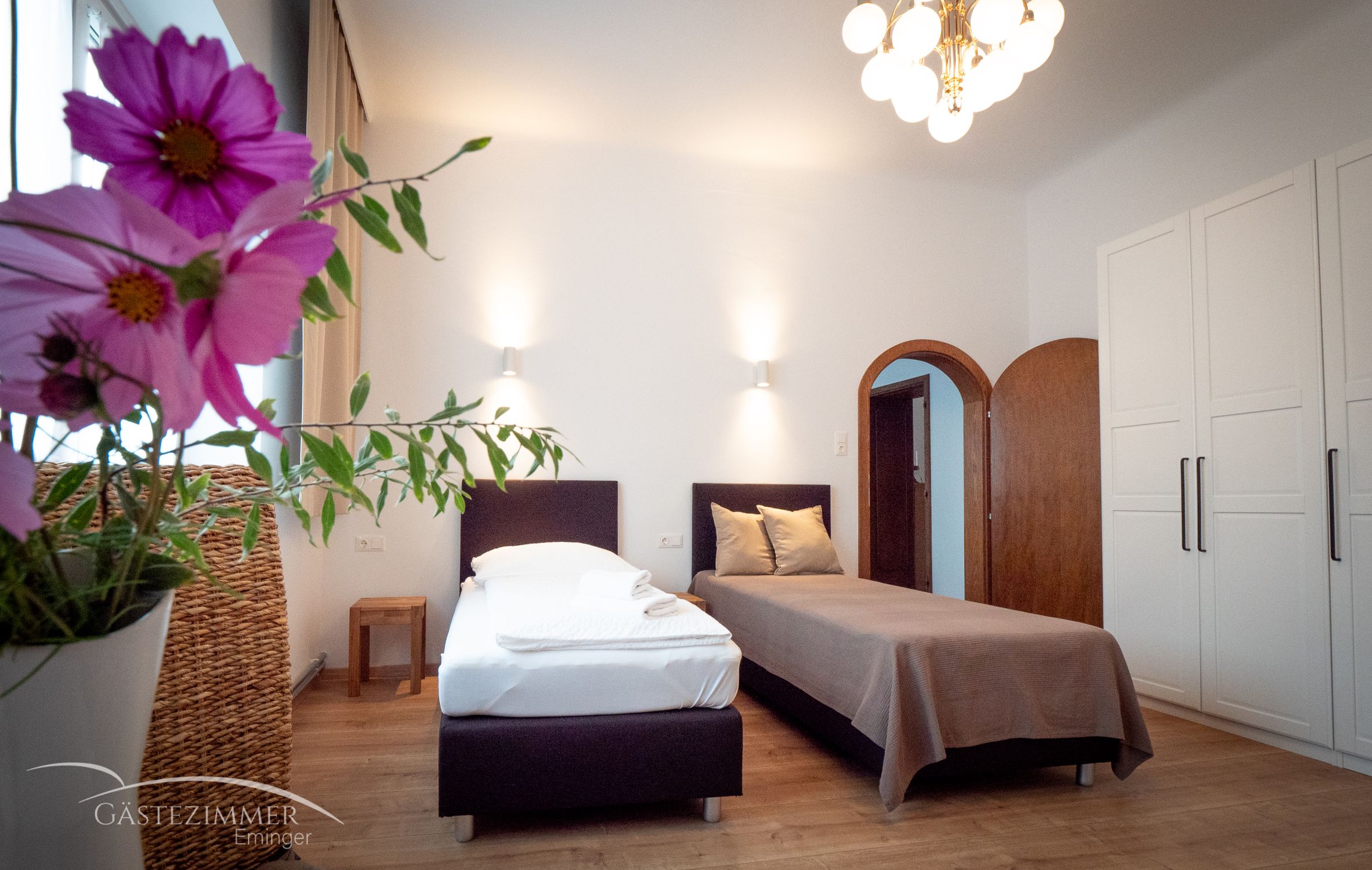 Útulný dvoulůžkový pokoj s květinami v popředí, dvěma postelemi, šatní skříní a lampou na stropě.