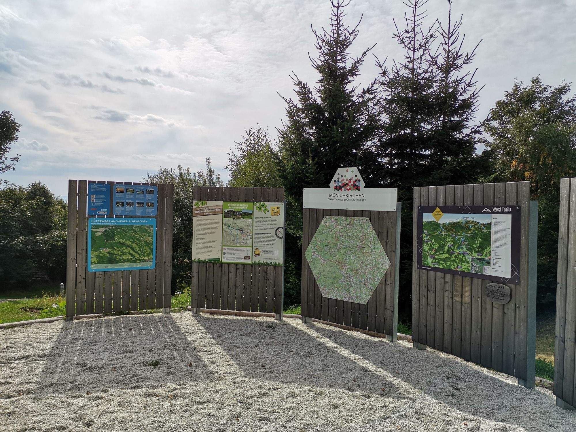Informační tabule s mapami a informacemi u výchozího bodu pro pěší turistiku v Mönichkirchenu, obklopené stromy.