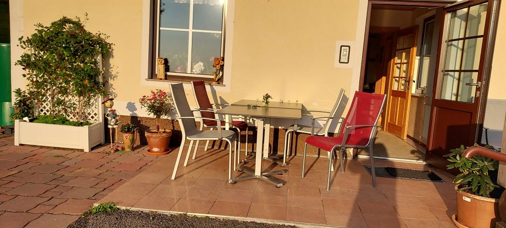 Terasa se stolem a židlemi před budovou, obklopená rostlinami.