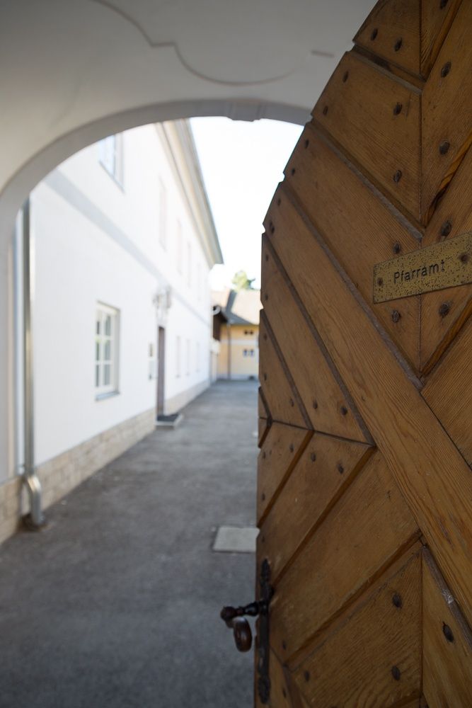 Dřevěná brána s nápisem "Pfarramt" ústí na nádvoří s bílými budovami.