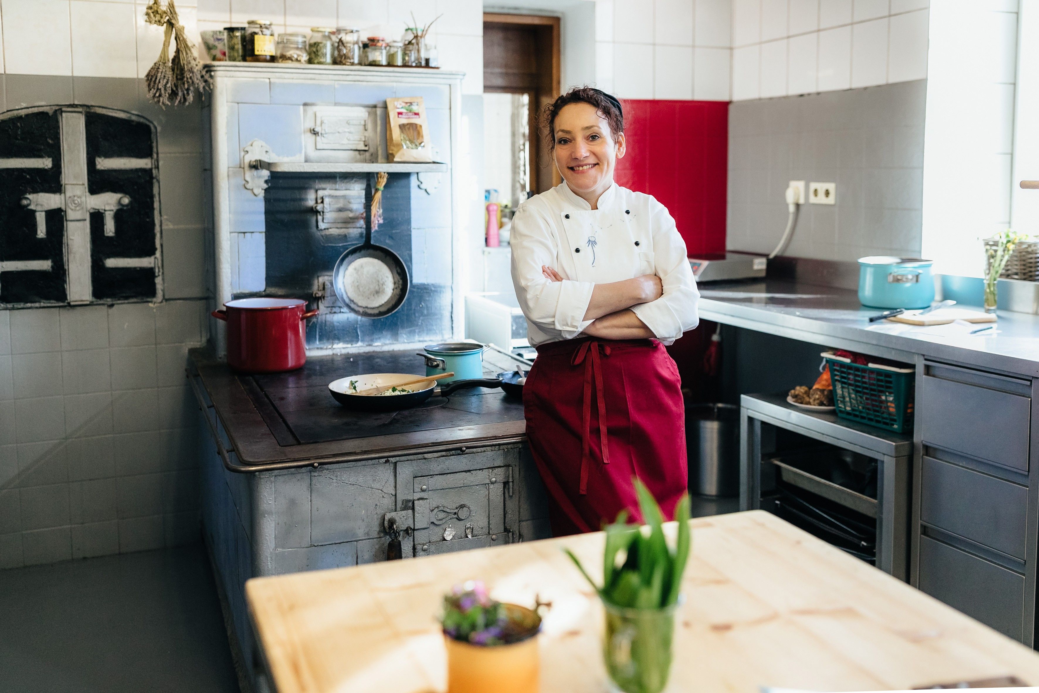 Kuchař v bílém saku a červené zástěře stojí v kuchyni před starým sporákem.