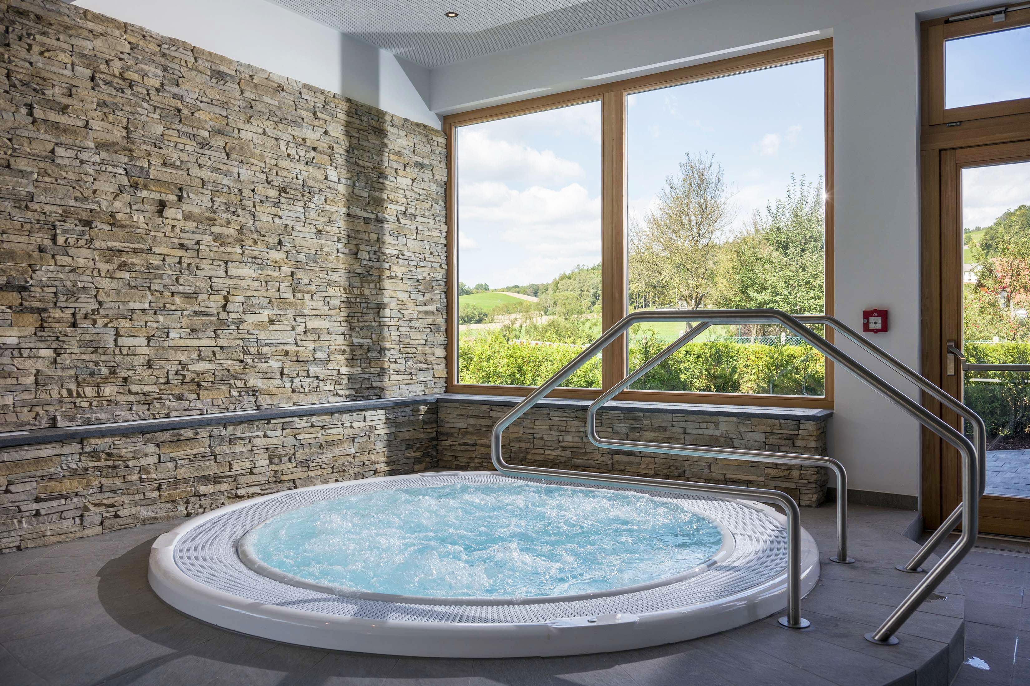 Wellness místnost s vířivkou, kamennými stěnami a velkým oknem s výhledem do zeleně.