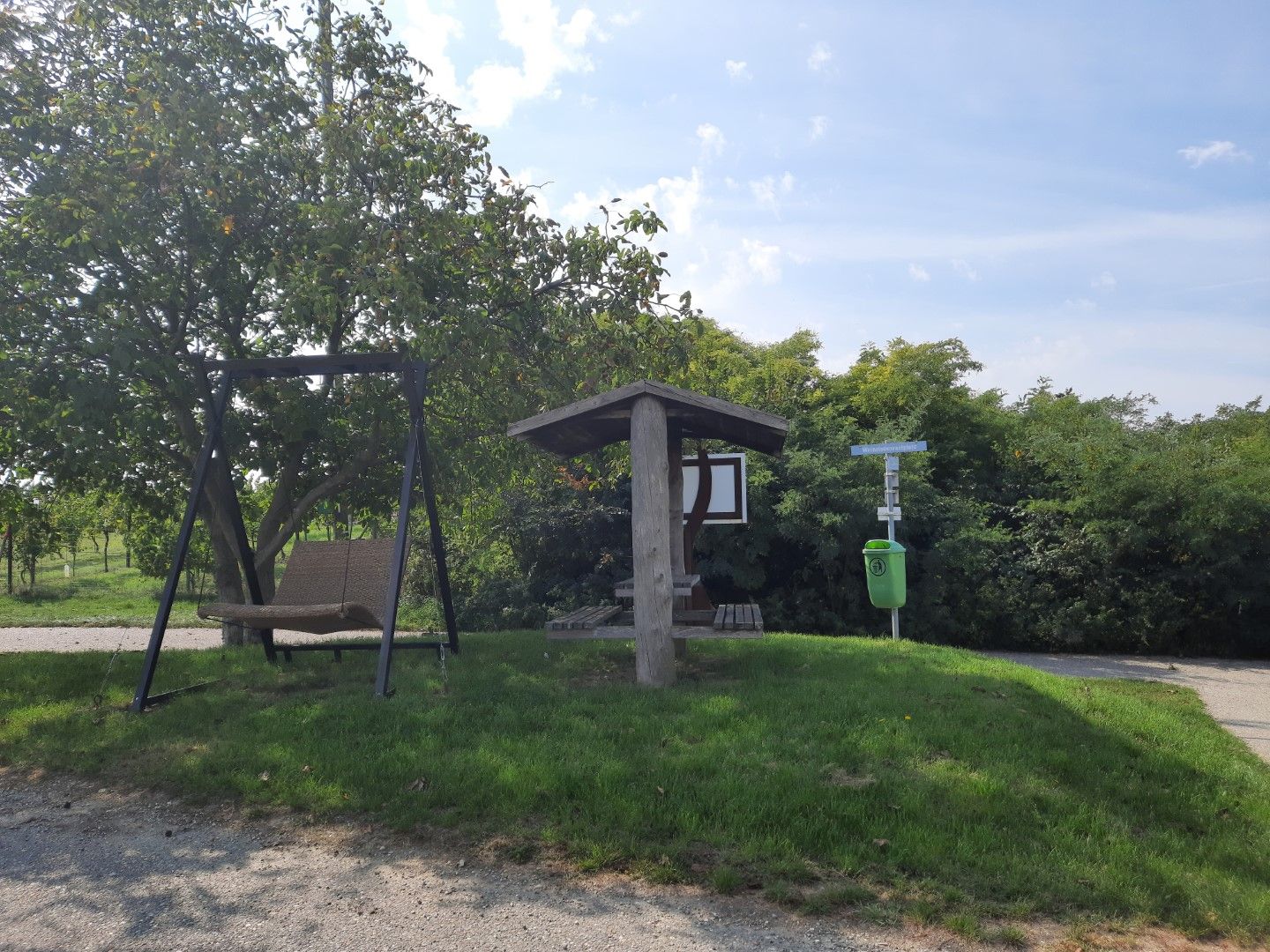 Odpočívadlo s houpačkou a zastřešeným posezením u stromu a odpadkového koše na louce.