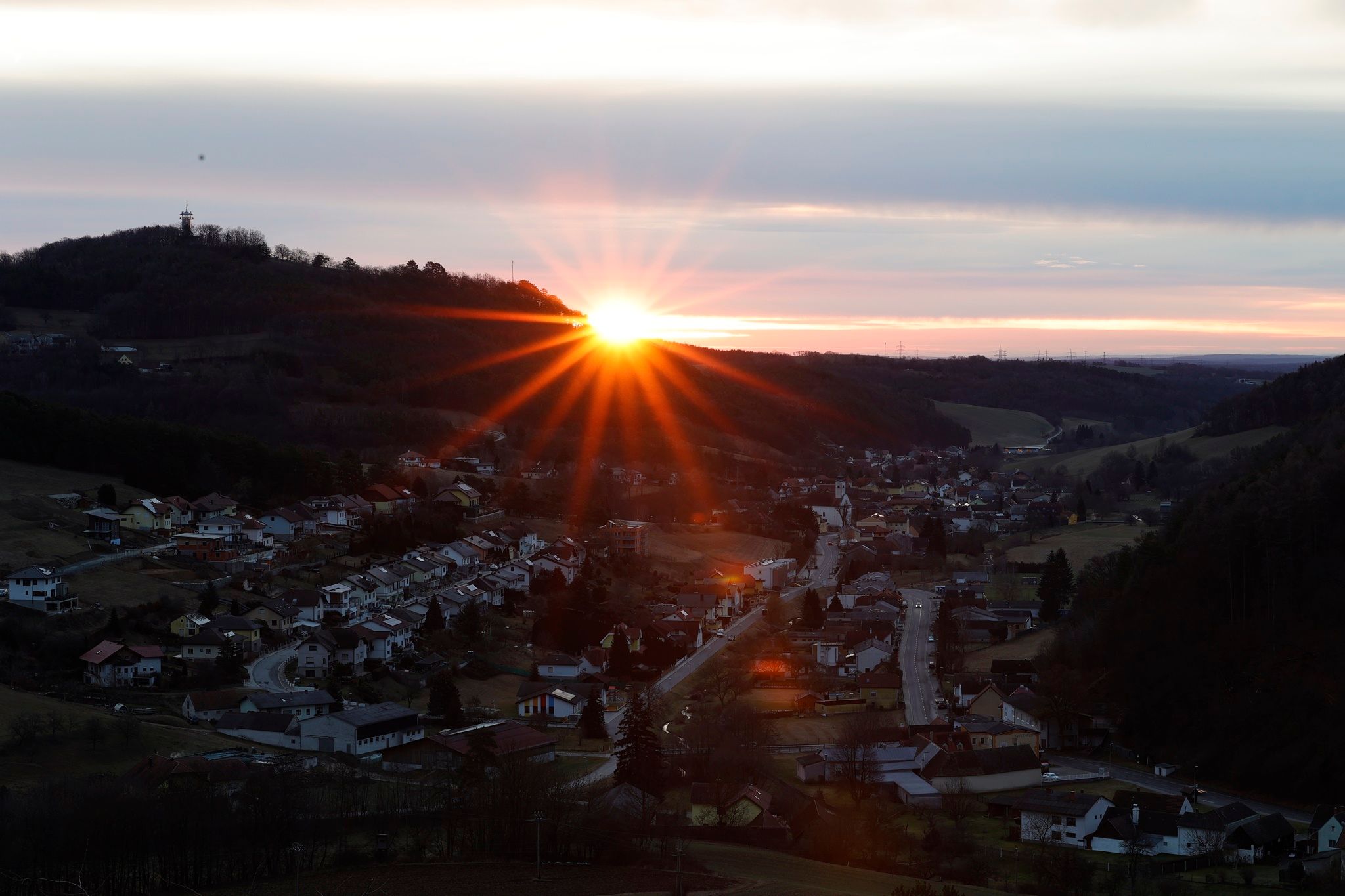 Východ slunce za kopcem s vesnicí v popředí.