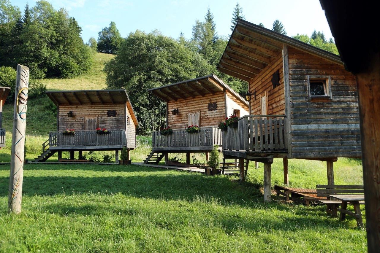 Tři dřevěné chatky na kůlech s verandami v zelené zalesněné oblasti.