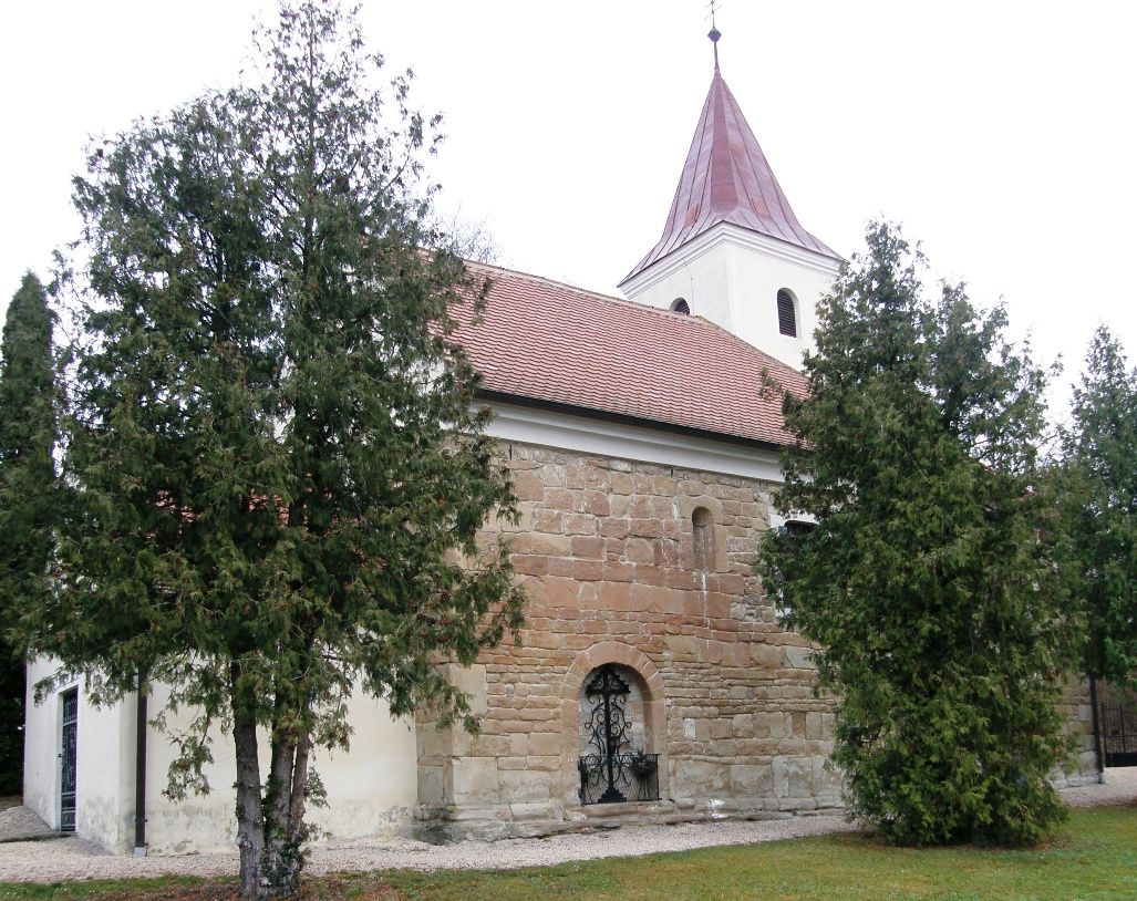 Starý kostel s červenou taškovou střechou a věží, obklopený stromy.