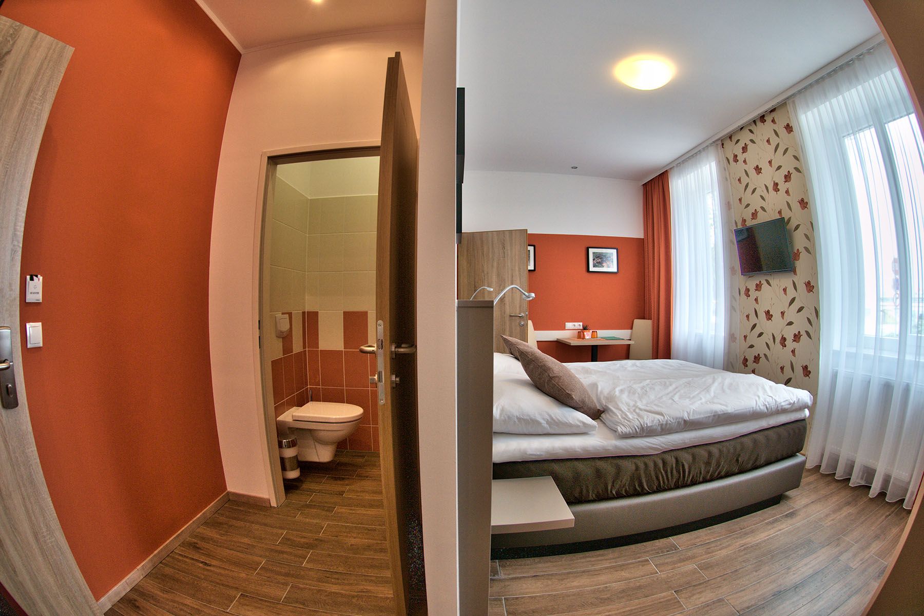 Moderní pokoj pro hosty s manželskou postelí, psacím stolem a televizí. Vedle je malá koupelna s WC.
