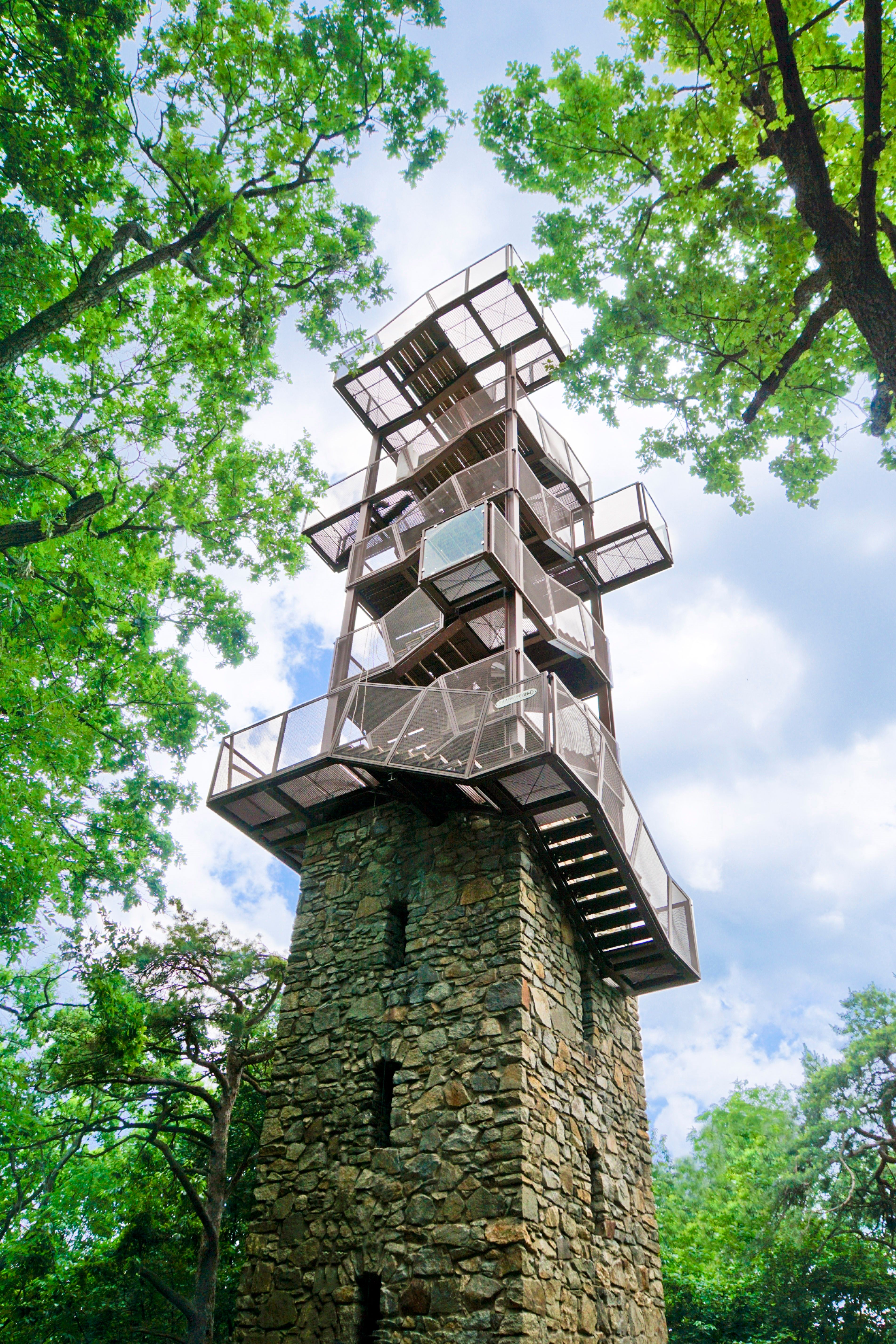 K modré obloze se tyčí moderní rozhledna z kovu a kamene, obklopená zelenými stromy.