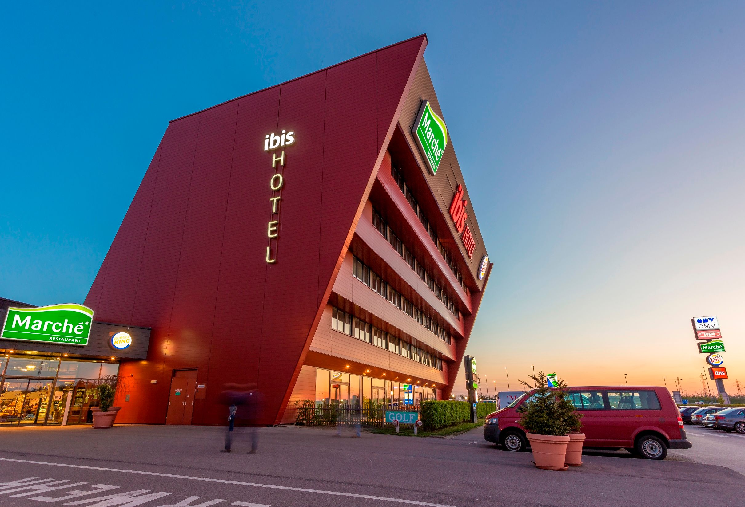 Moderní hotelová budova se šikmou fasádou a osvětleným nápisem "ibis HOTEL".