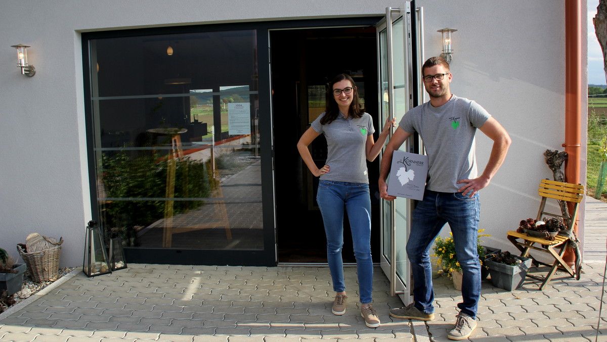 Před budovou s prosklenými dveřmi stojí dvě osoby, obě mají na sobě šedá trička a džíny. Jedna osoba drží ceduli s nápisem "Kornherr".