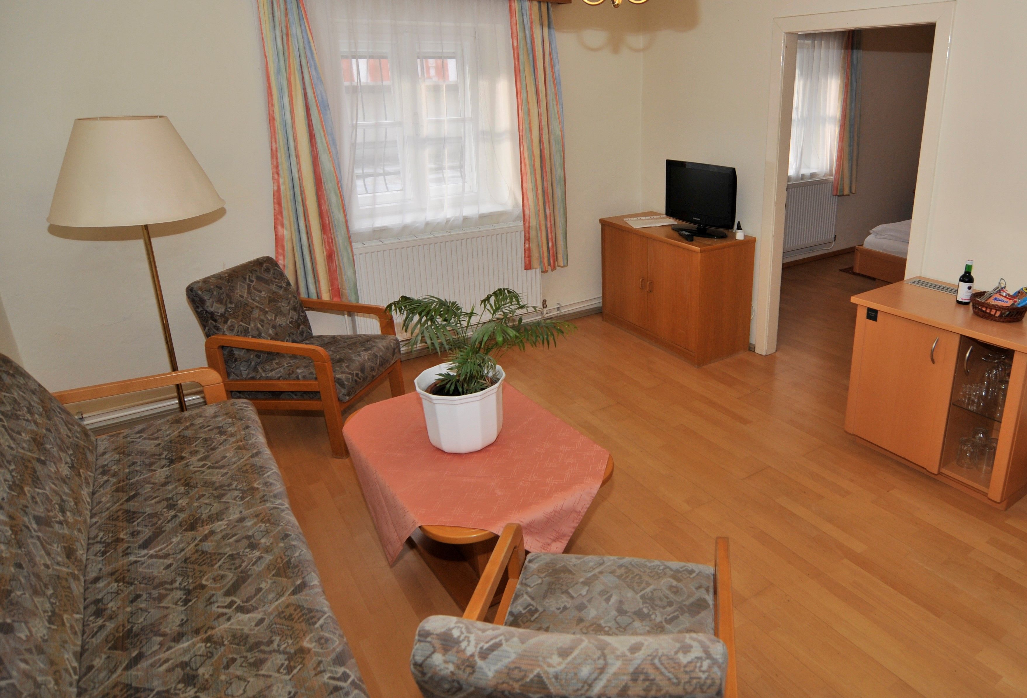 Obývací pokoj s pohovkou, dvěma křesly, stolem s rostlinami, TV a přístupem do ložnice.