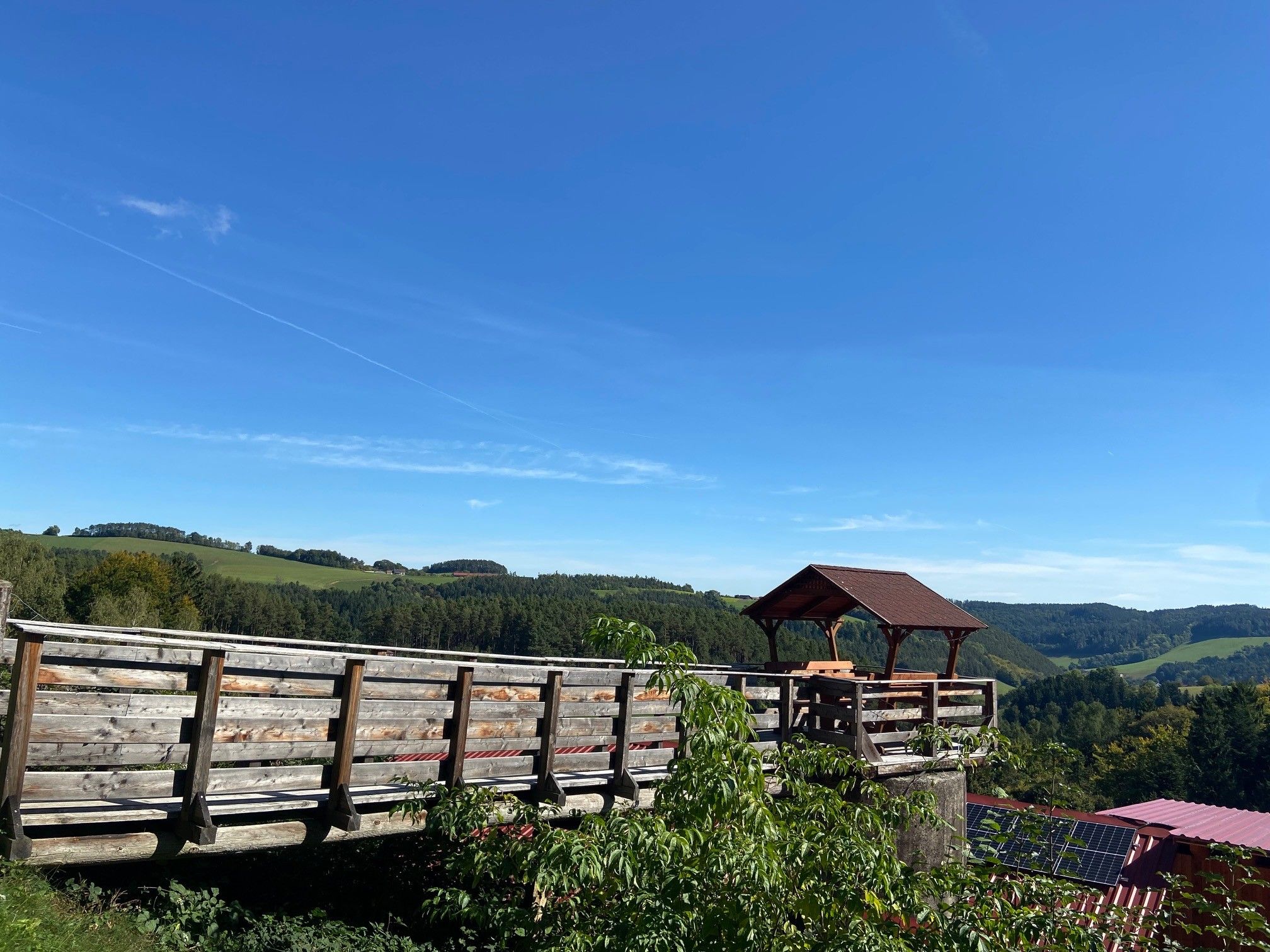 Dřevěná plošina s pavilonem a výhledem na zelené kopce a modrou oblohu.