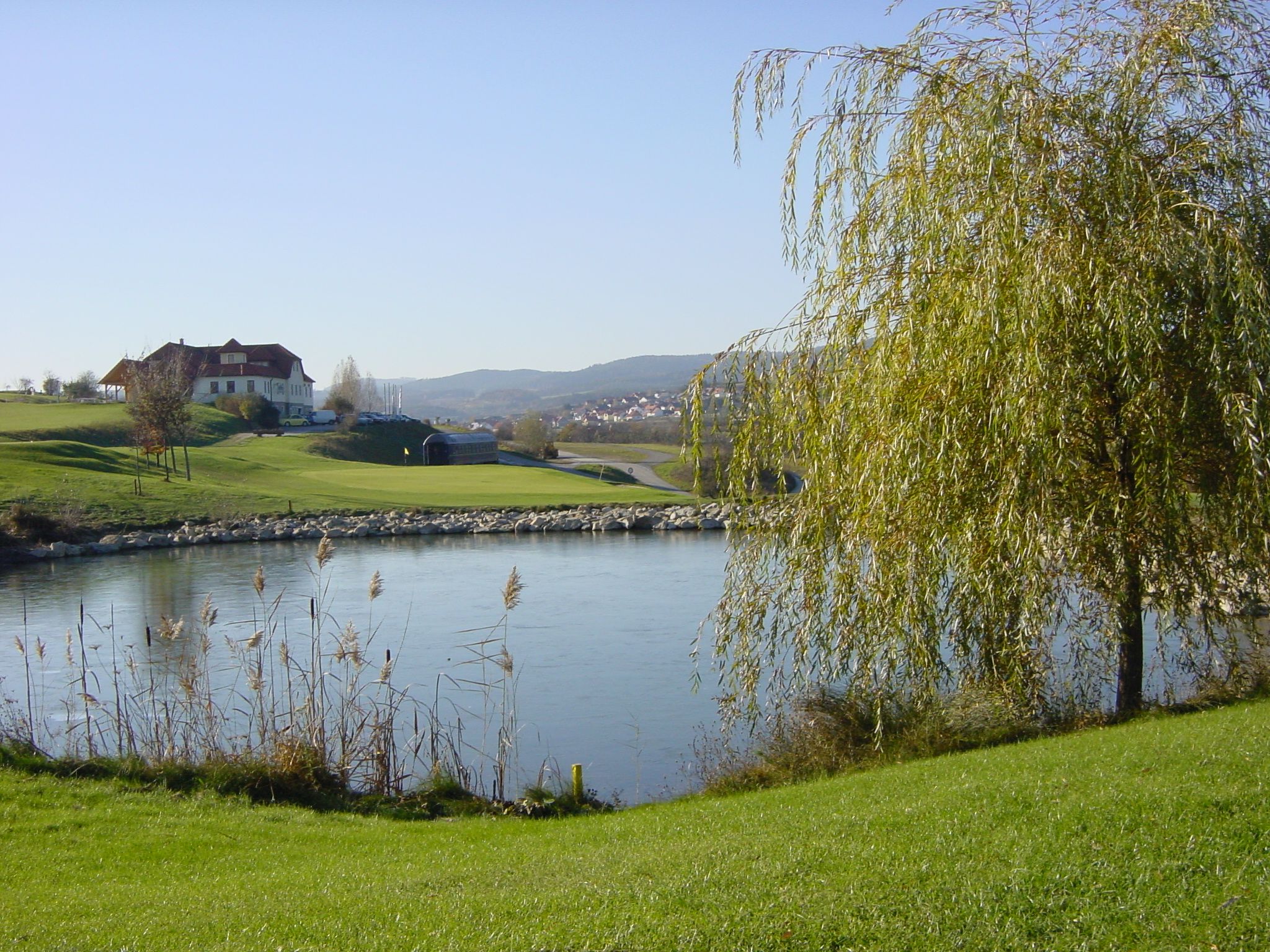 Krajina s golfovým hřištěm, rybníkem a budovami v pozadí.