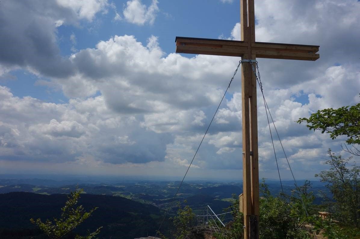 Vrcholový kříž na vyhlídkové hoře Burgsteinmauer s výhledem na zataženou oblohu a krajinu.