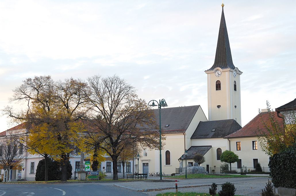 Kostel v Absdorfu s hodinovou věží a okolními budovami, v popředí stromy.