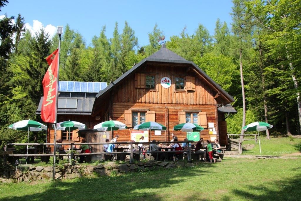 Dřevěná chata v lese se slunečníky a lidmi na terase.