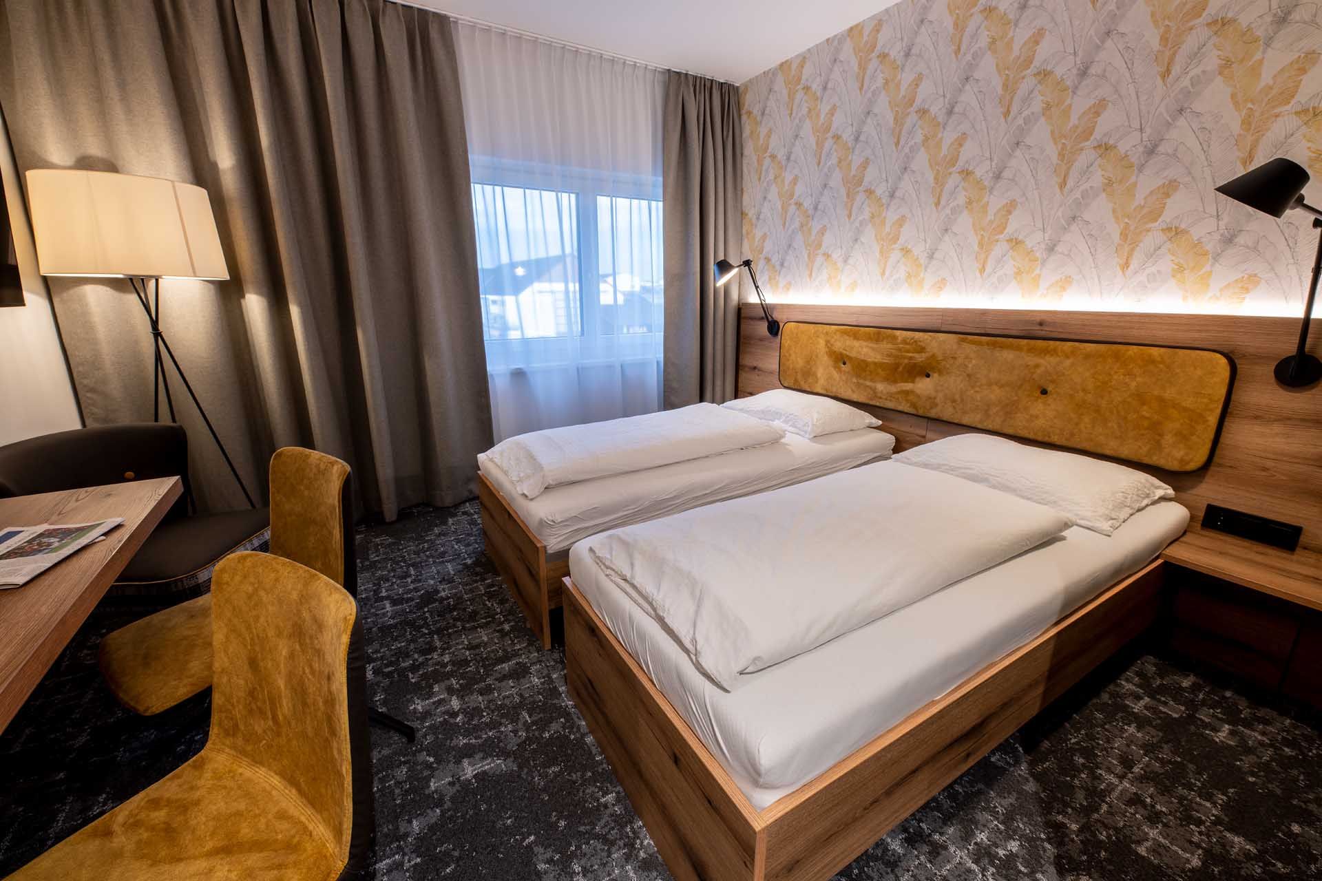 Moderní pokoj pro hosty se dvěma samostatnými lůžky, dřevěným nábytkem, žlutými židlemi a stojací lampou. Světlé závěsy a vzorované tapety na stěnách.