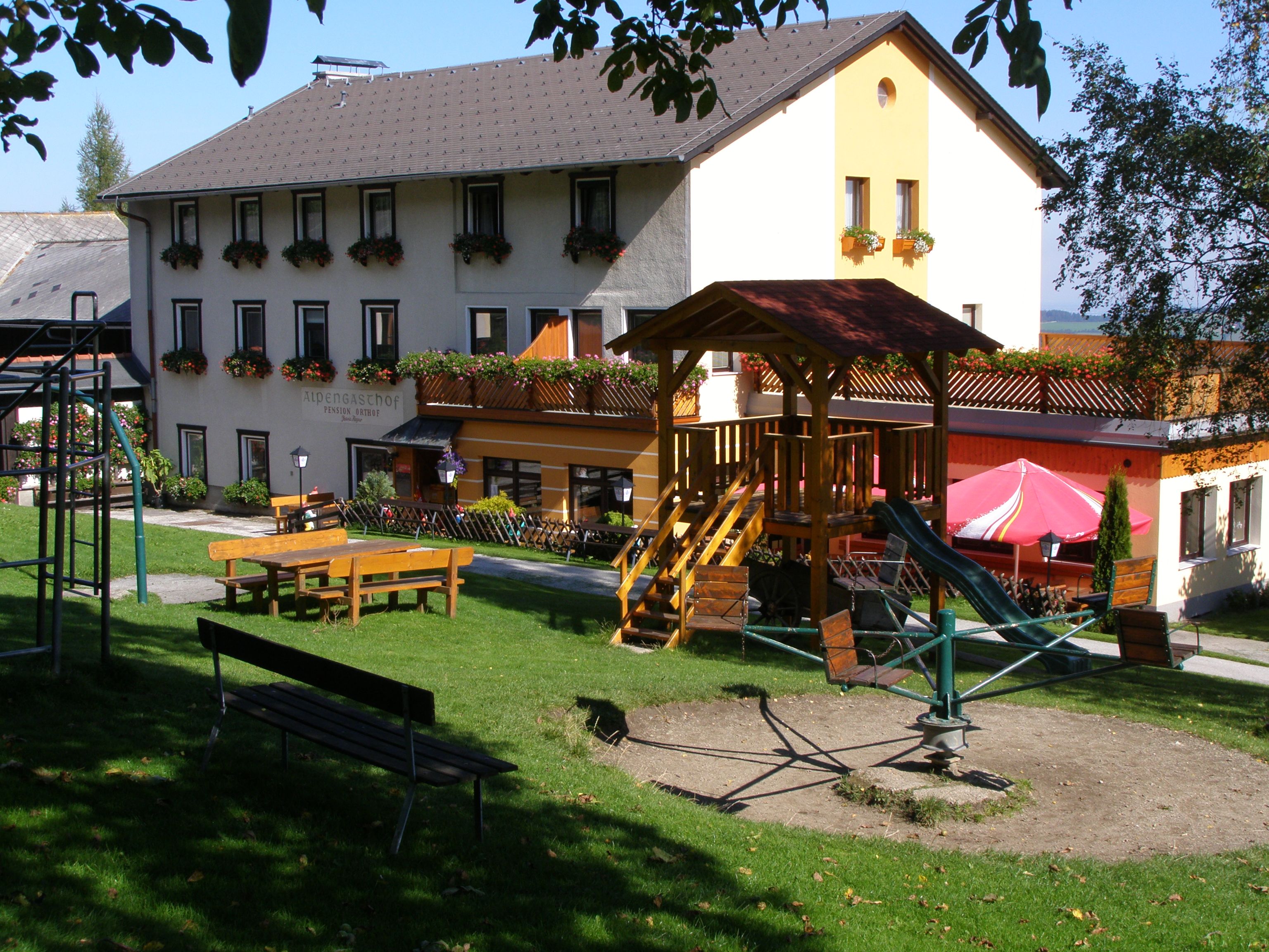 Hostinec s hřištěm v popředí, obklopený zelenými loukami a stromy.