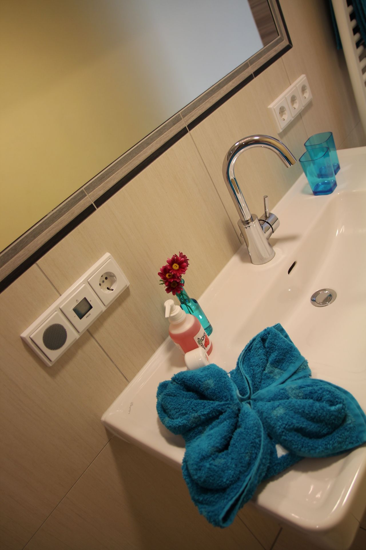Umyvadlo s modrým ručníkem, dávkovačem mýdla a vázou s květinami.