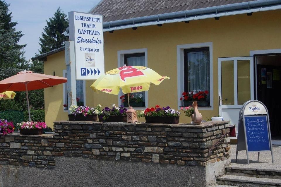 Pohled na žlutou budovu s nápisy pro potraviny, trafiku a hostinec. Před budovou jsou slunečníky a květiny.